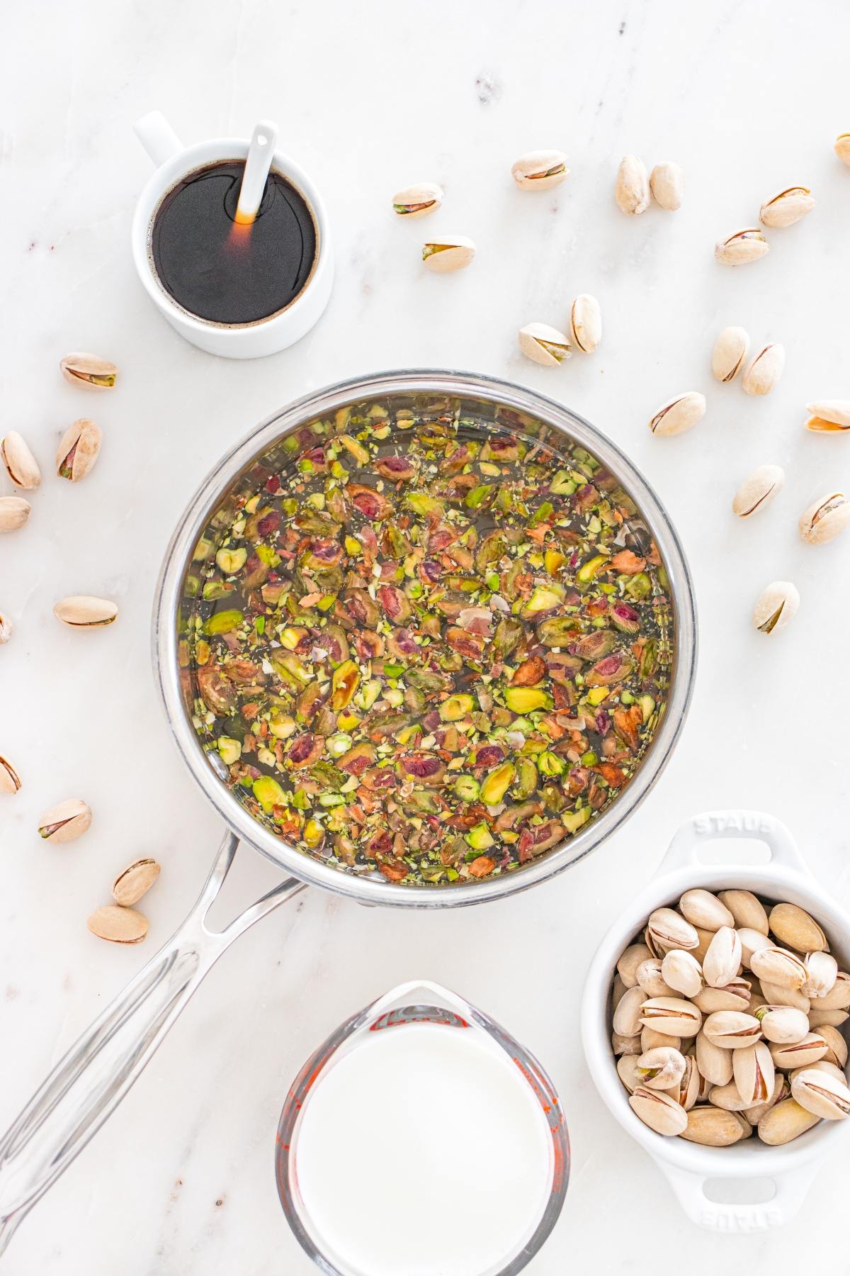A saucepan with chopped pistachios, surrounded by milk, pistachios, and a cup of dark liquid on a white surface.