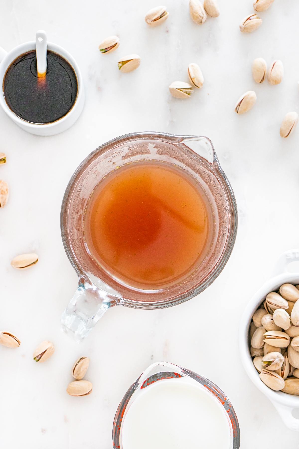 A glass measuring cup of brown liquid surrounded by pistachios, milk, and a small cup of dark syrup.