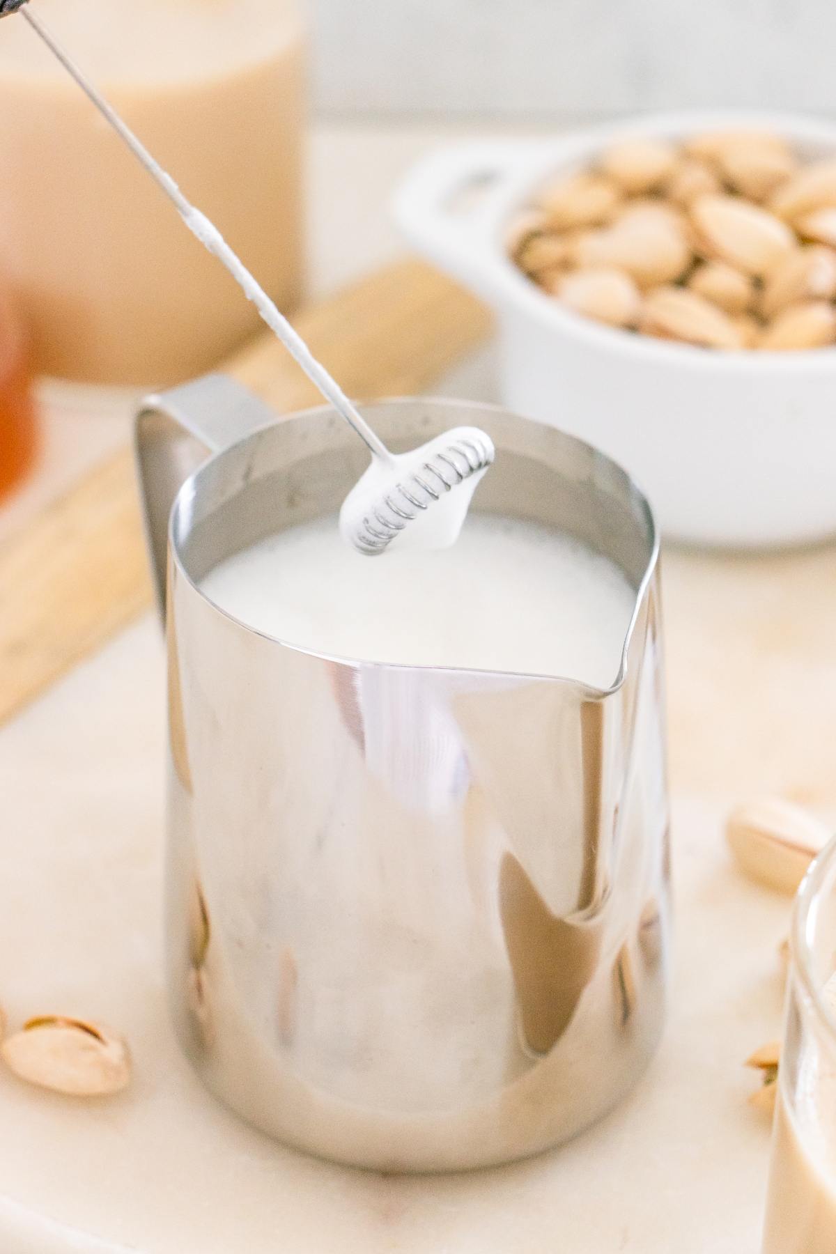 A milk frother whipping milk in a metal pitcher, with a bowl of pistachios in the background.