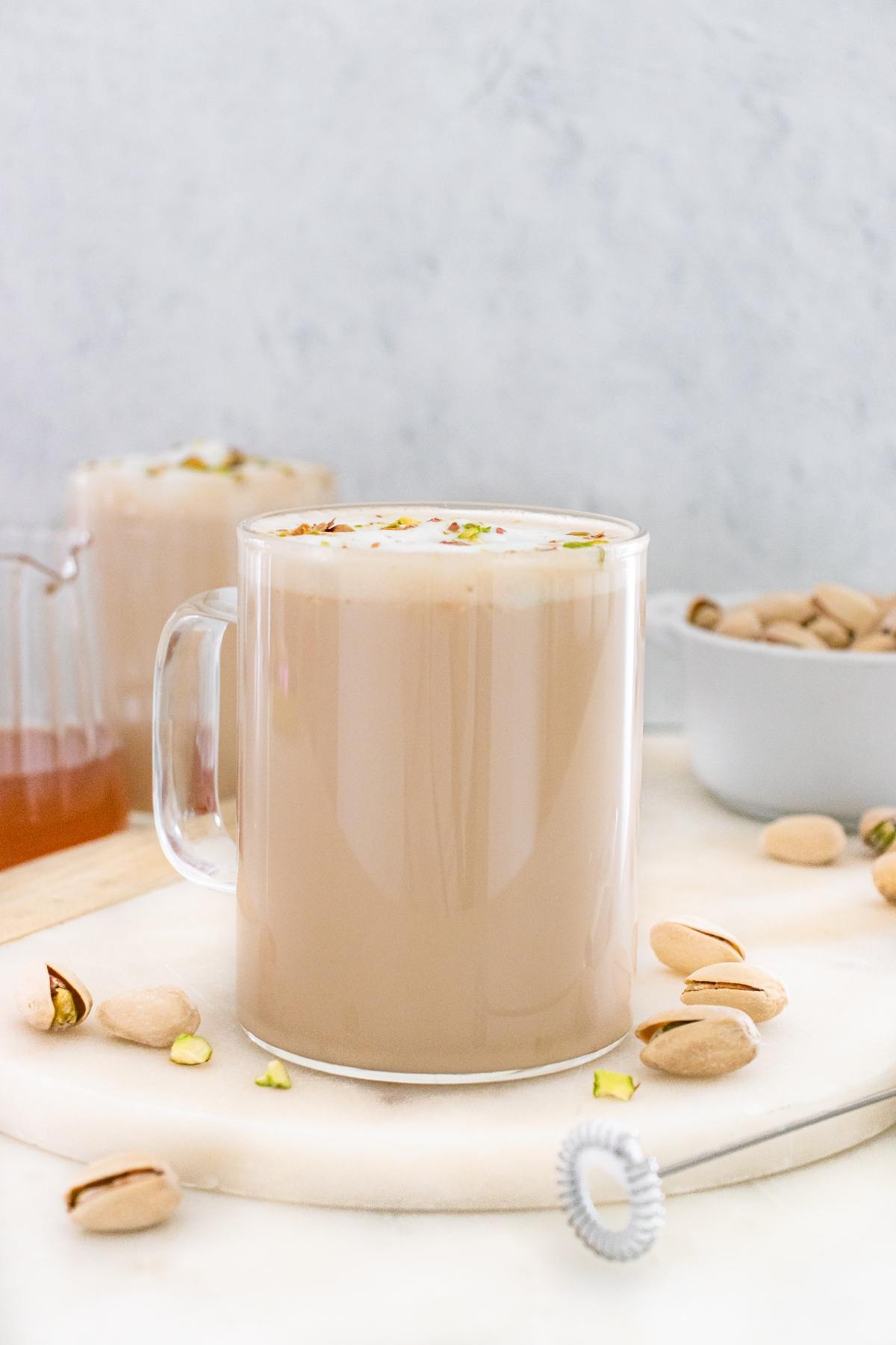 A glass mug of pistachio latte on a marble board, with scattered pistachios and a bowl in the background.