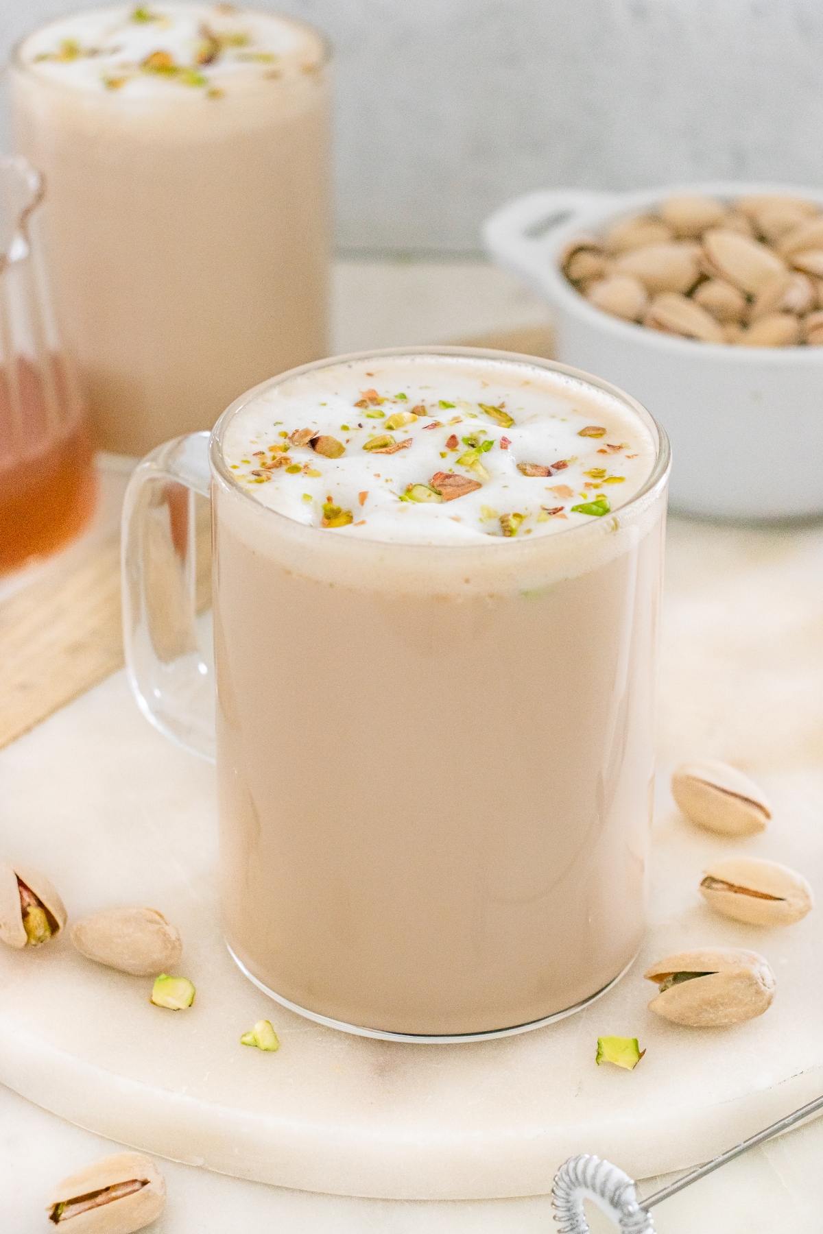 A glass mug of chai topped with frothed milk and chopped pistachios, with pistachios scattered around.