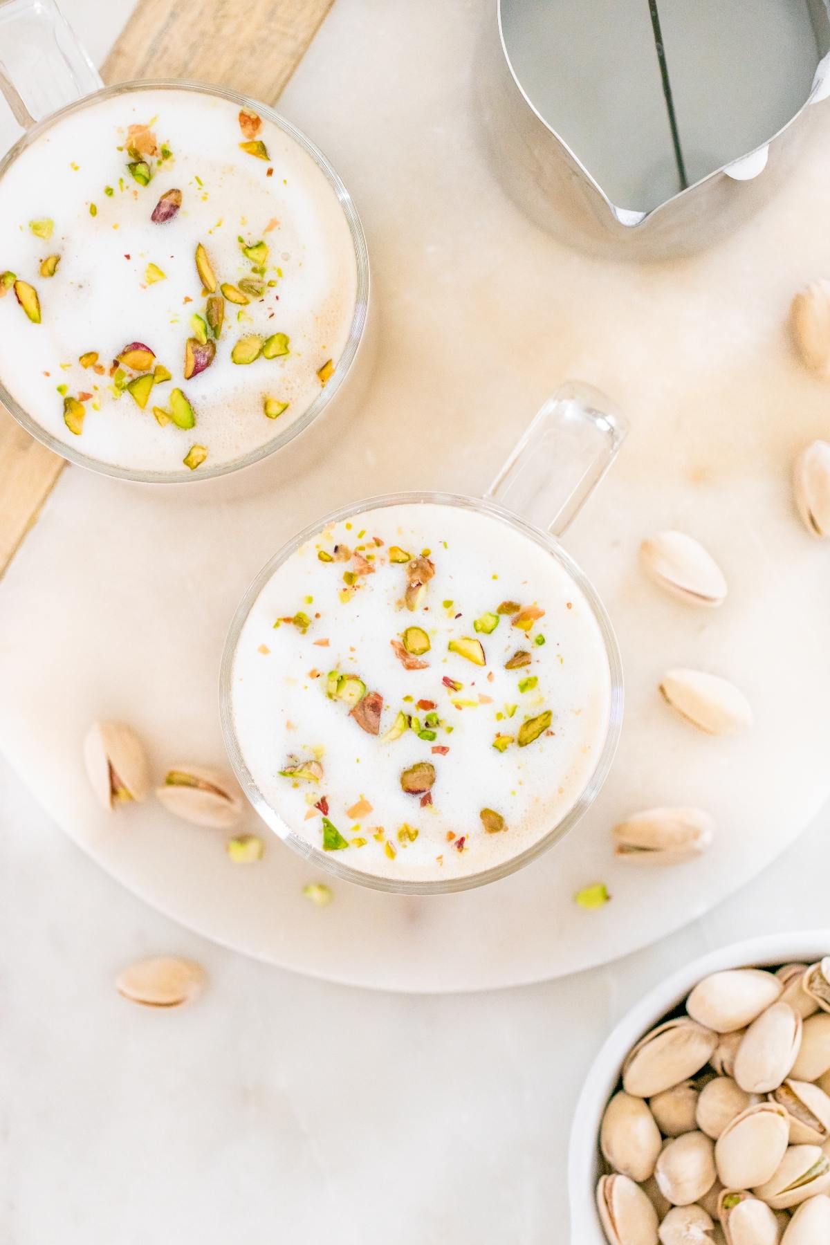 Two cups of frothy pistachio latte topped with chopped pistachios, surrounded by whole pistachios on a table.