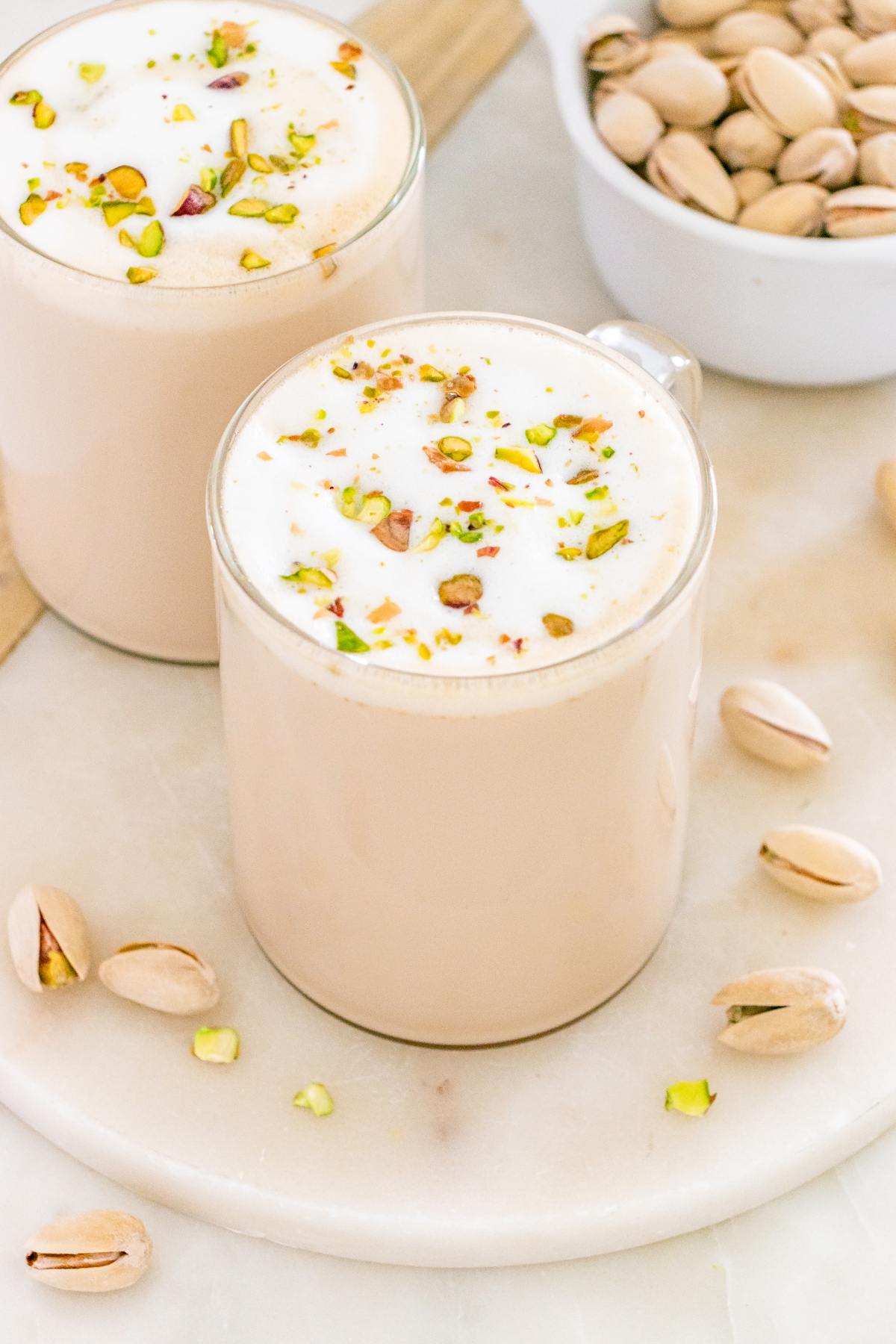 Two frothy pistachio lattes in glass mugs, topped with chopped pistachios, beside a bowl of pistachios.