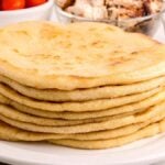 A stack of fresh pita bread on a white plate with bowls of vegetables in the background.