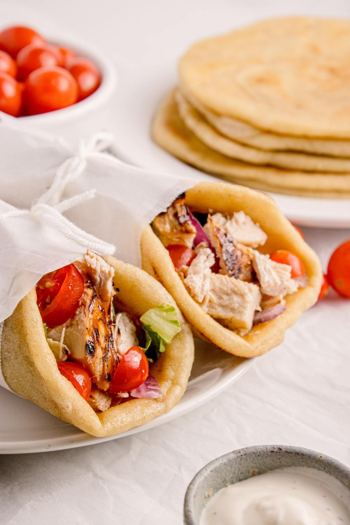 Two pita wraps filled with grilled chicken, lettuce, tomatoes, and onions, with flatbreads and tomatoes in background.
