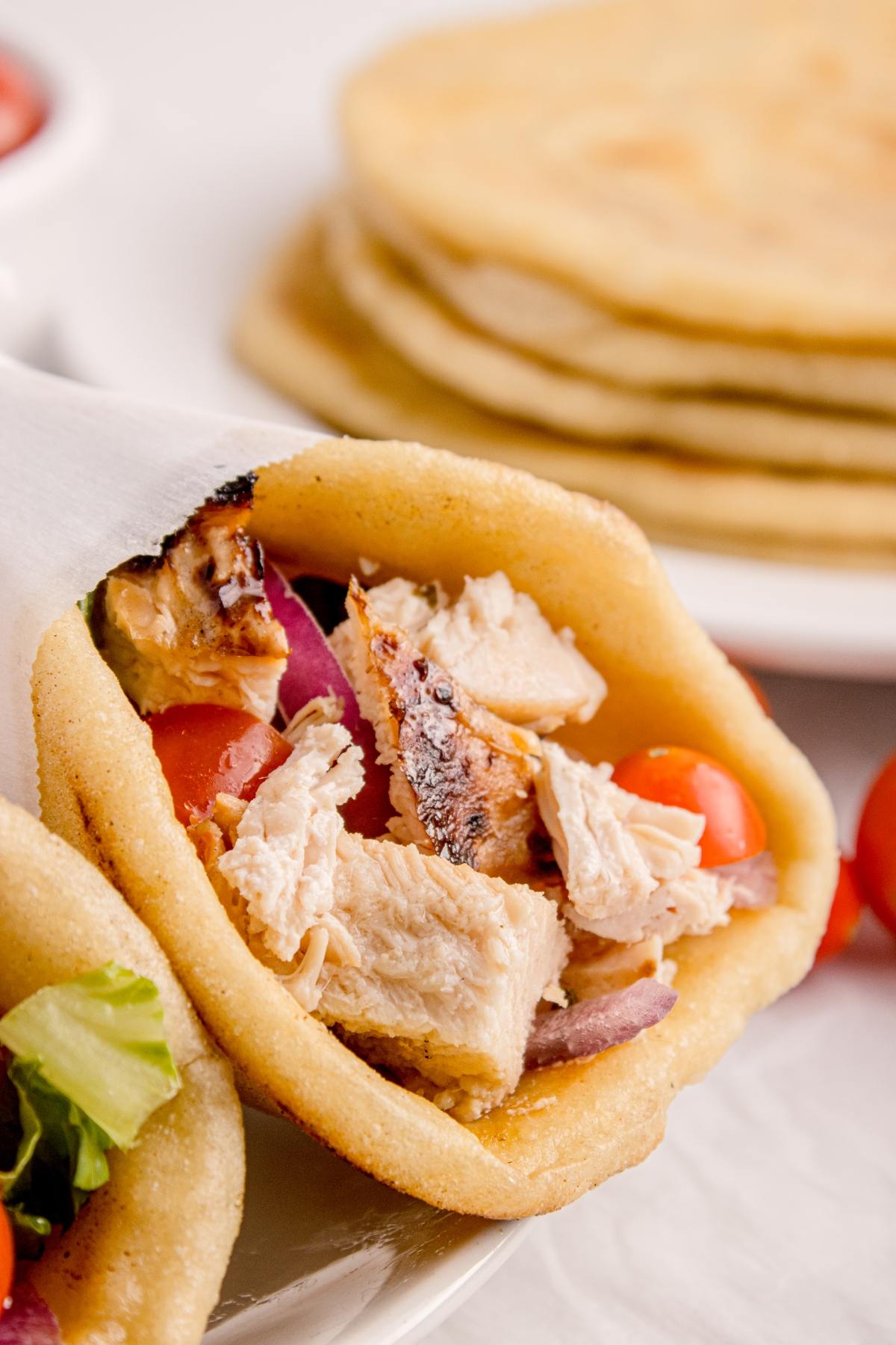 Close-up of a pita stuffed with grilled chicken, tomatoes, and onions, with more pitas stacked in the background.