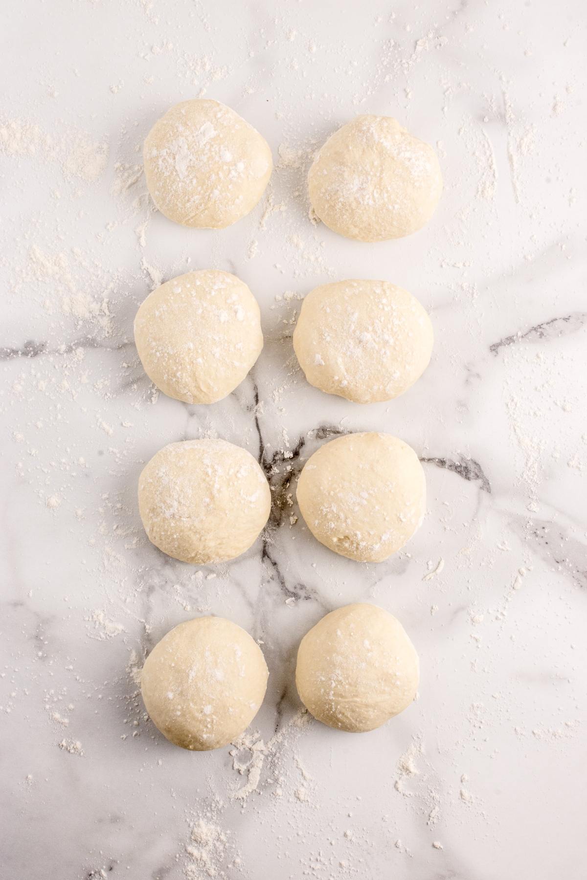 Eight balls of dough dusted with flour are arranged in two vertical rows on a marble surface.