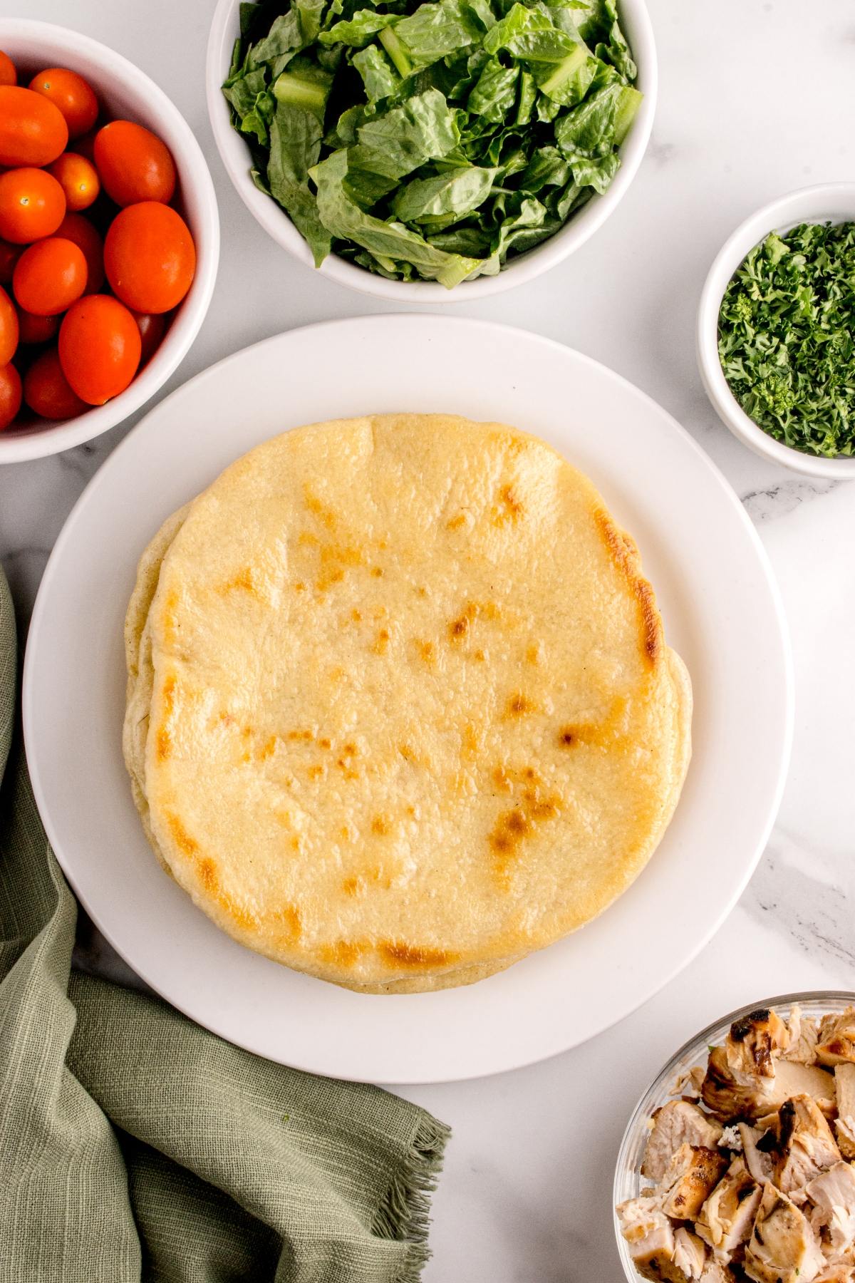 A plate of pita bread surrounded by bowls of lettuce, tomatoes, parsley, and chopped grilled chicken.