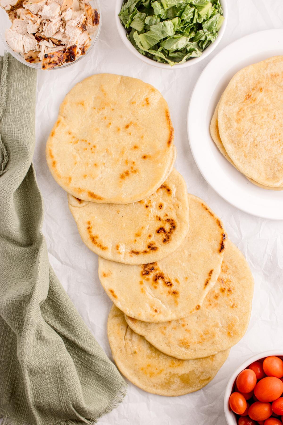Five pieces of flatbread with bowls of lettuce, shredded chicken, and tomatoes on a white surface.