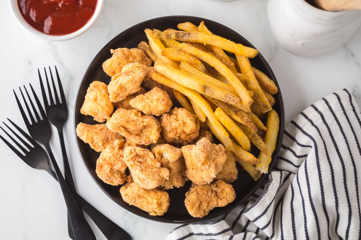A plate of chicken nuggets, popcorn chicken, and French fries with a bowl of ketchup and black forks nearby on a marble surface.