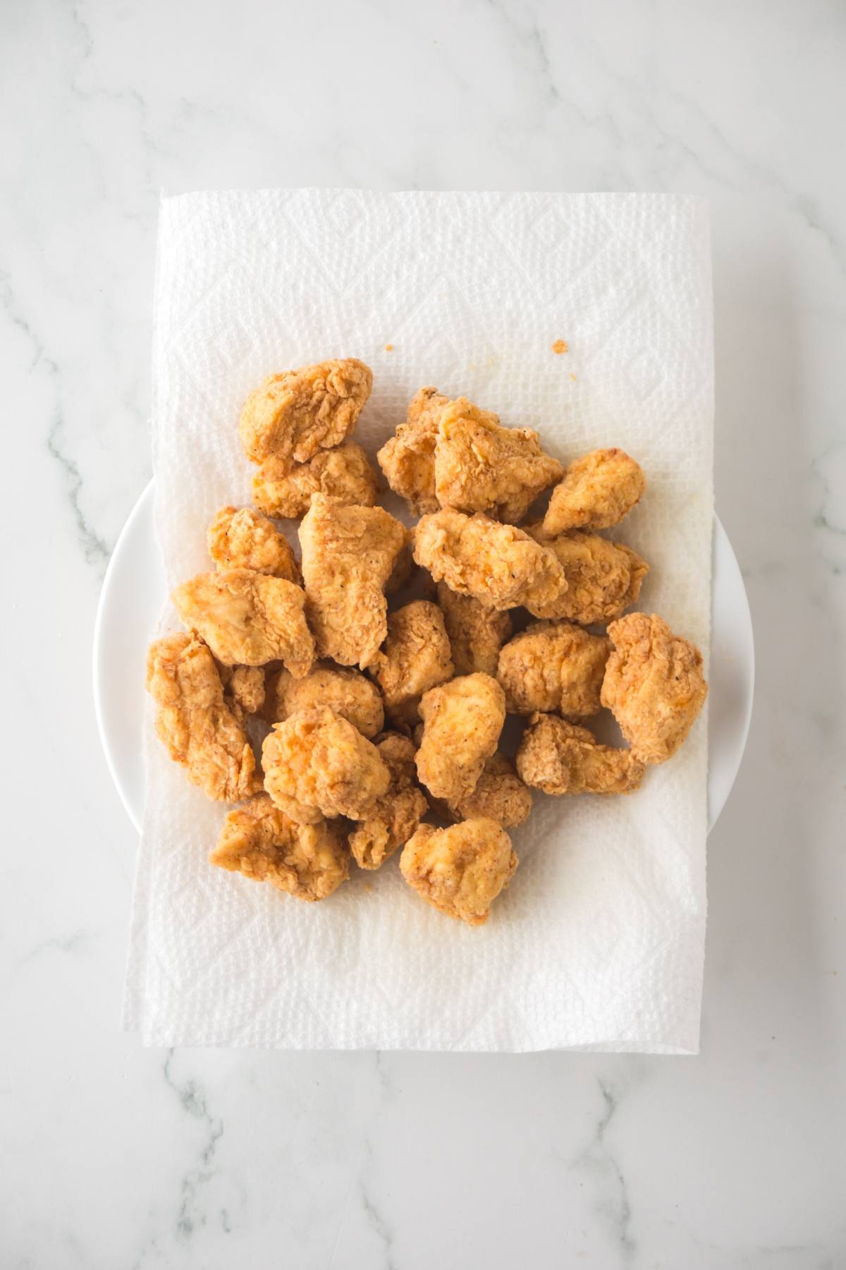 Golden fried chicken pieces on a paper towel-lined plate, viewed from above on a marble surface.