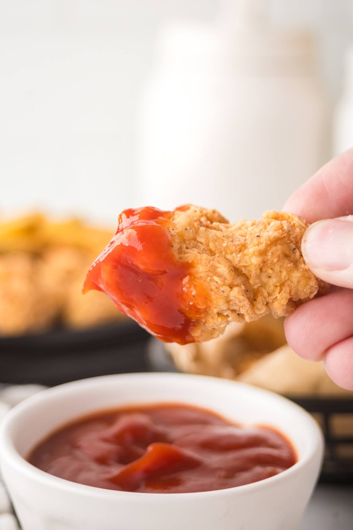 A hand dips a crispy chicken tender into a bowl of red ketchup sauce.