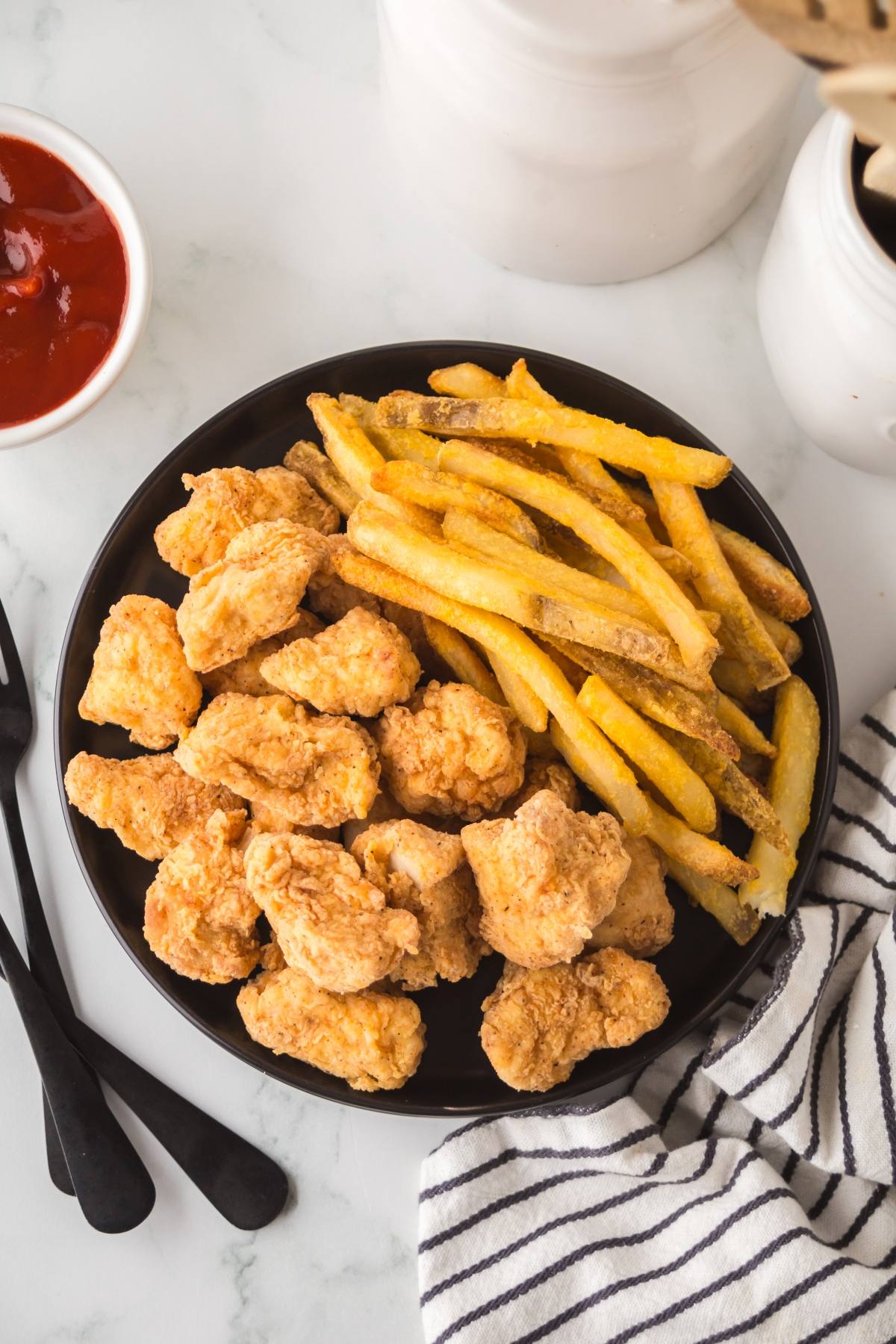 A black plate with fried chicken nuggets and French fries, next to ketchup and a striped napkin.