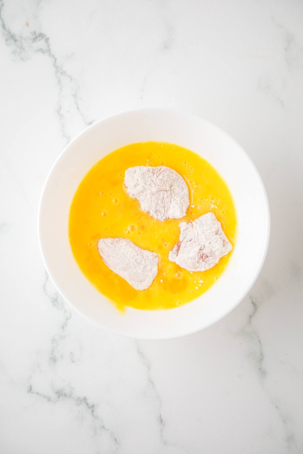 Three flour-coated chicken pieces sit in beaten eggs in a white bowl on a marble surface.