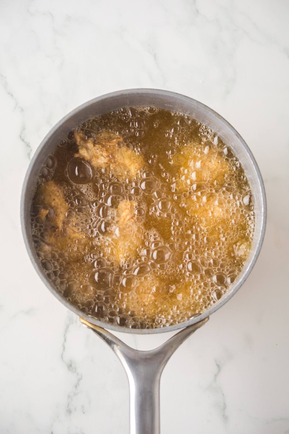 Chicken pieces are frying in bubbling oil in a saucepan on a white marble surface.