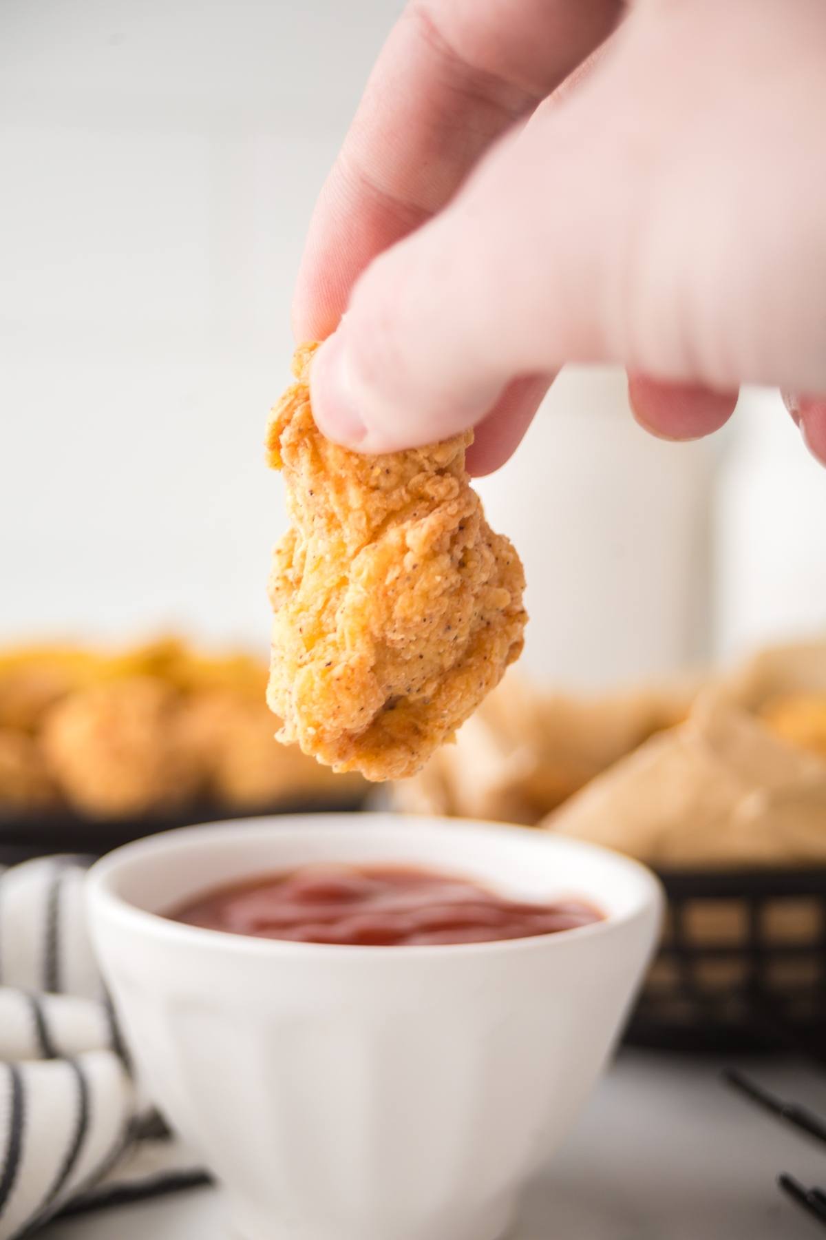 A hand holds a piece of fried chicken above a bowl of ketchup, ready for dipping.