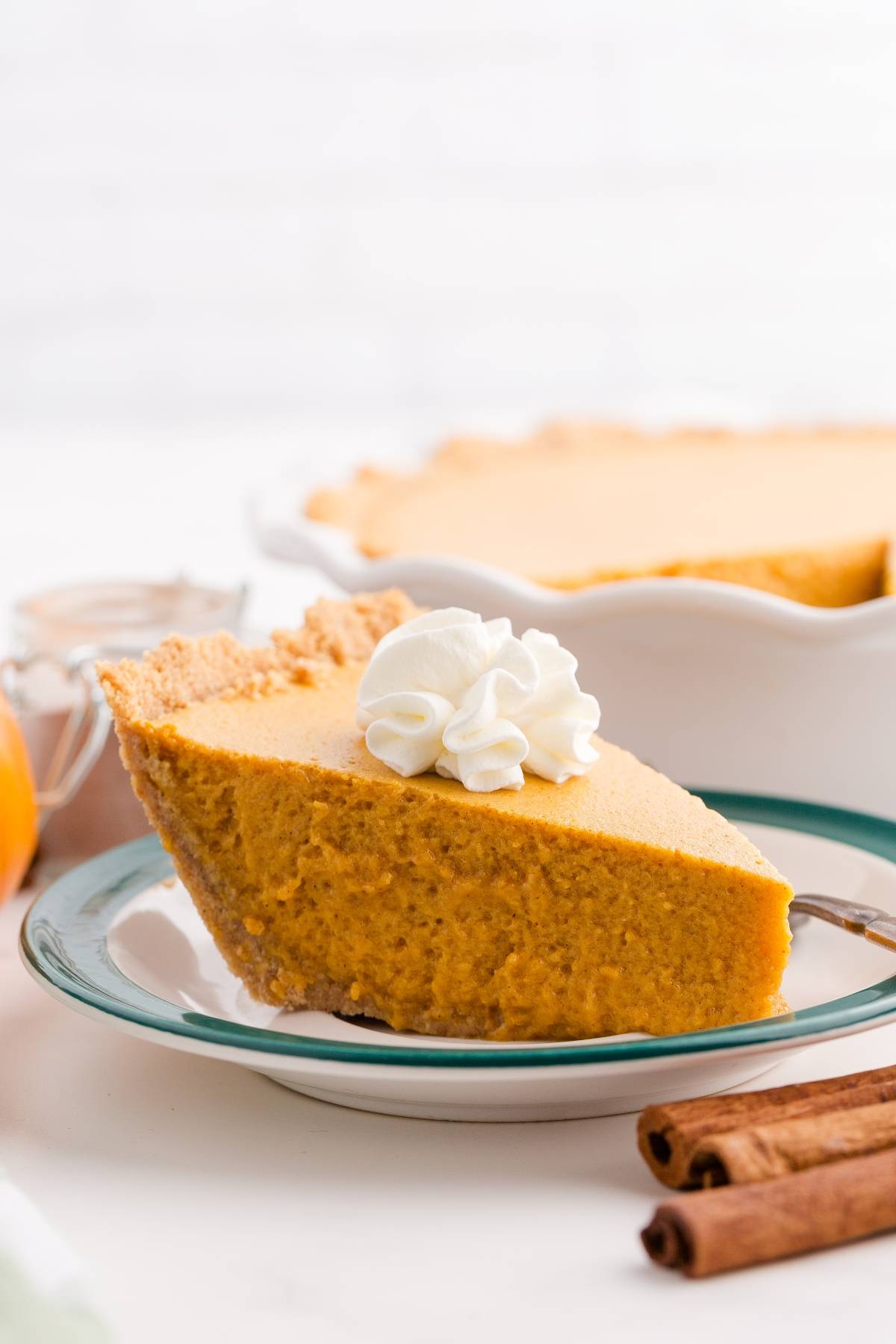 A slice of pumpkin pie with whipped cream on top, served on a plate with cinnamon sticks nearby.