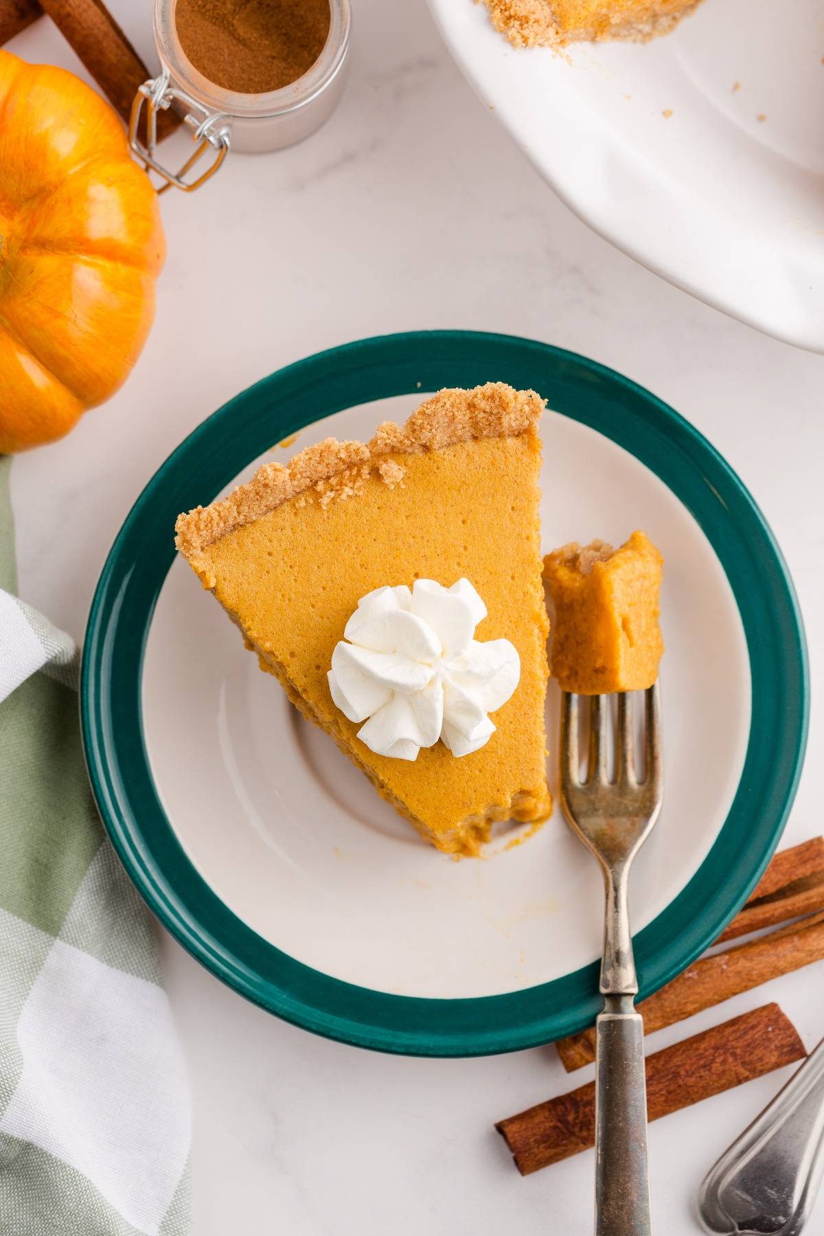 A slice of pumpkin pie with whipped cream on a plate, a fork with a bite, and cinnamon sticks nearby.