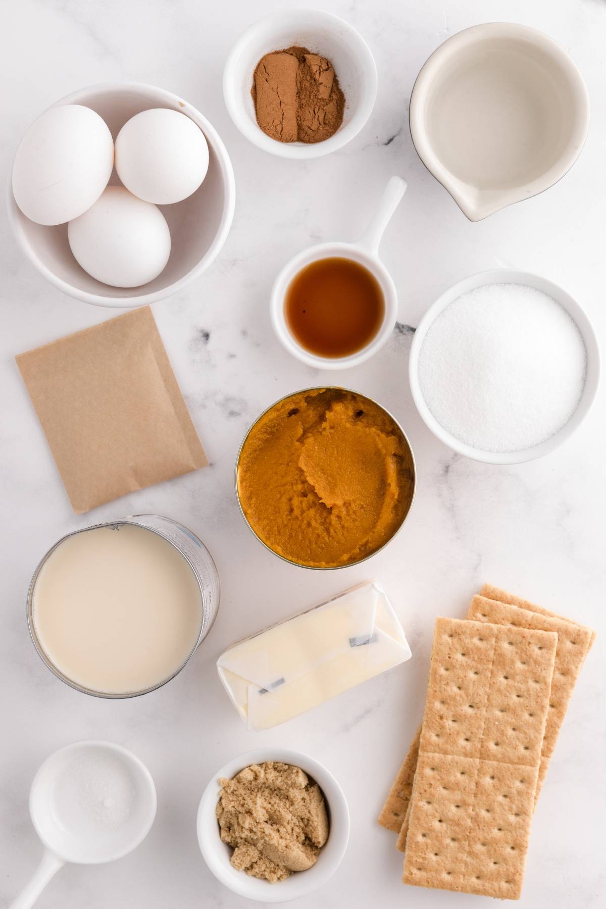Baking ingredients arranged on a white surface, including eggs, spices, pumpkin puree, sugar, and graham crackers.