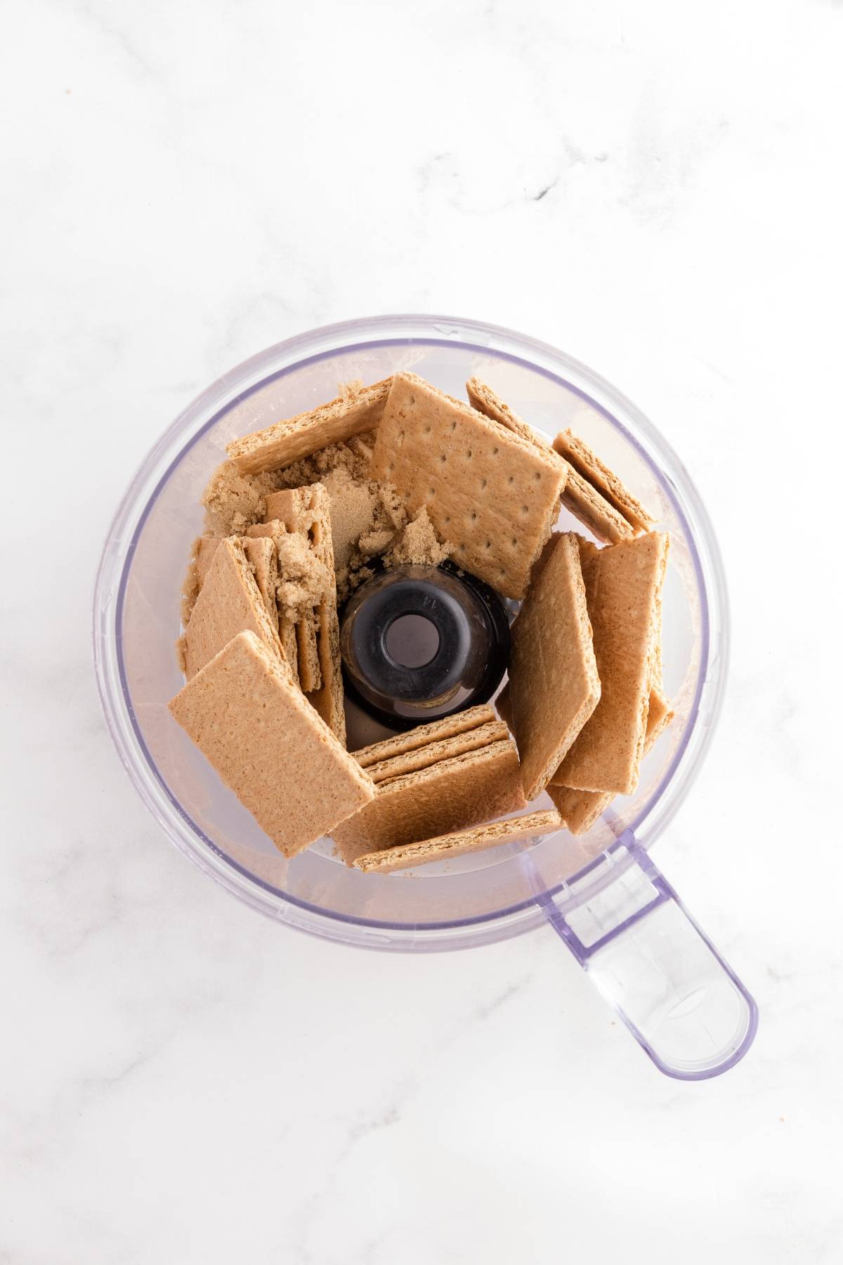 Overhead view of graham crackers and brown sugar in a food processor on a white surface.