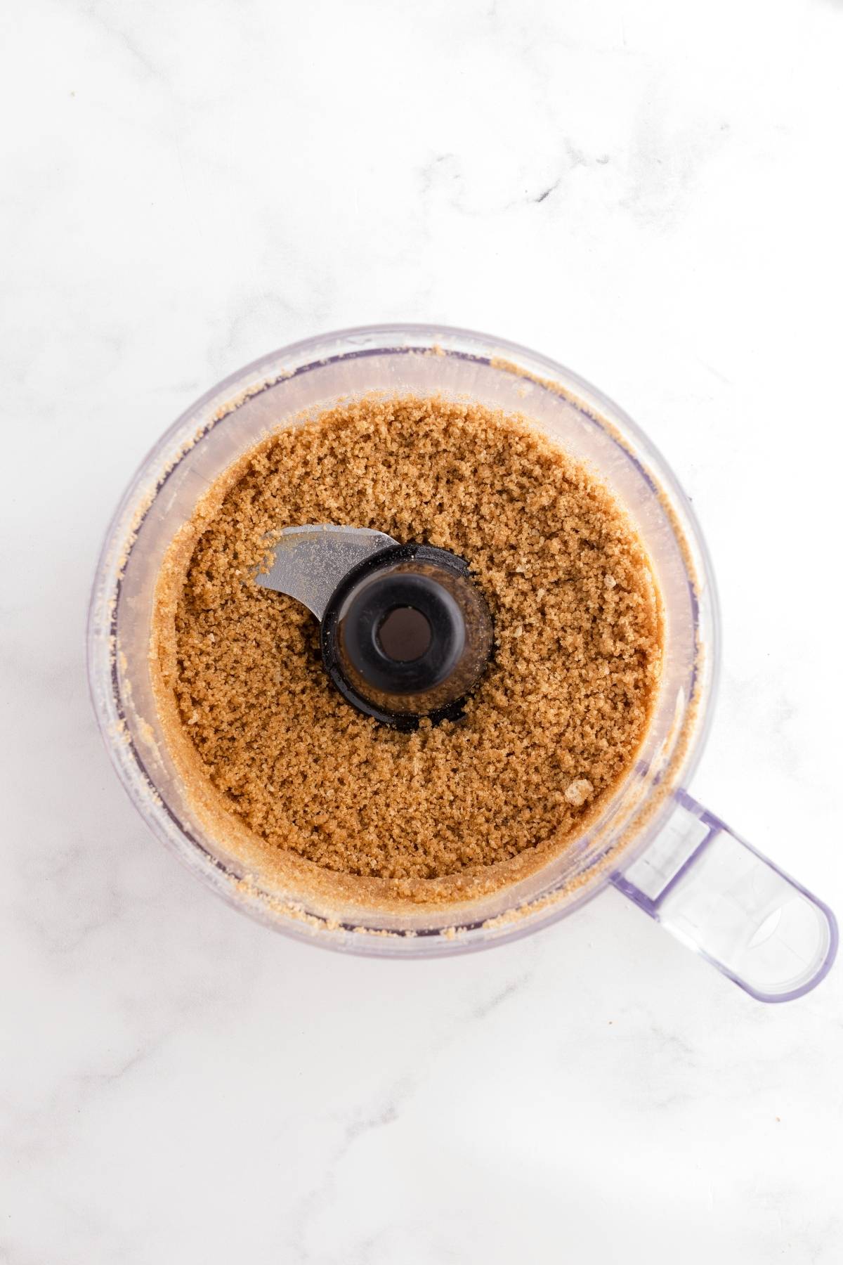 A food processor bowl filled with finely ground crumbs on a white surface.
