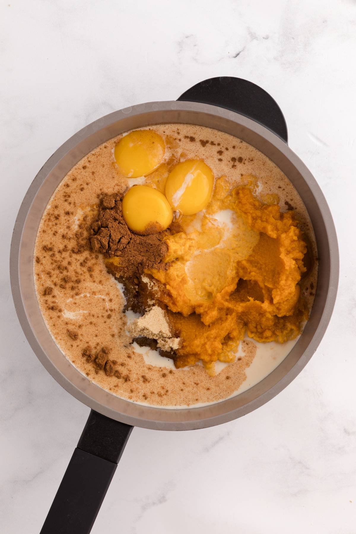 A saucepan with eggs, pumpkin puree, spices, sugar, and milk, viewed from above on a white surface.