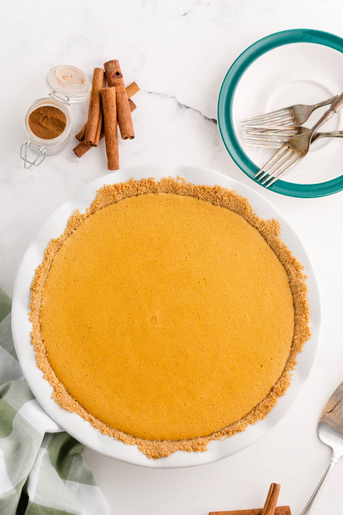 Pumpkin pie with a graham cracker crust on a marble surface, next to forks, plates, cinnamon sticks, and a jar of spice.