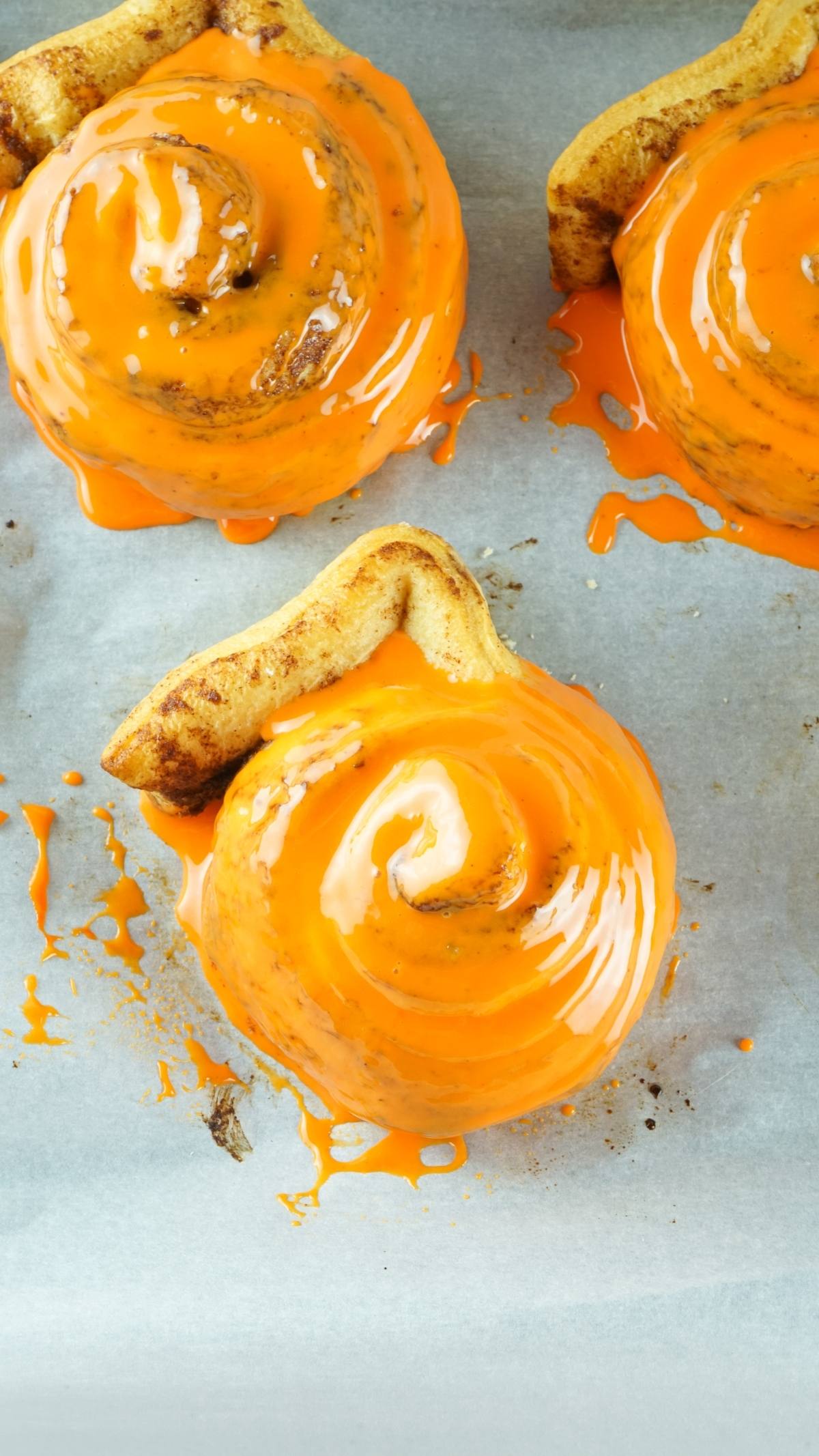 Three Pumpkin Shaped Cinnamon Rolls topped with bright orange glaze on parchment paper—an easy canned cinnamon roll hack for festive homemade pumpkin treats.