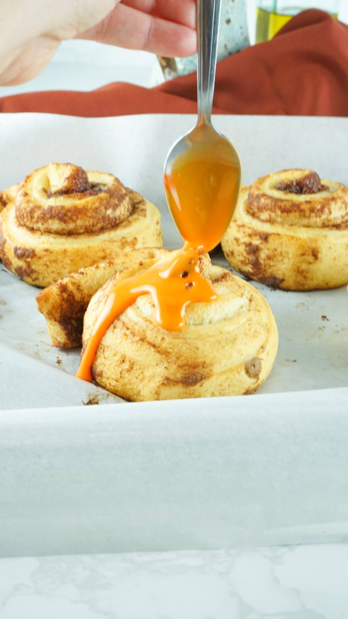 A spoon drizzles orange icing onto a cinnamon roll on a parchment-lined baking tray—a simple canned cinnamon roll hack perfect for an easy pumpkin shaped dessert idea.