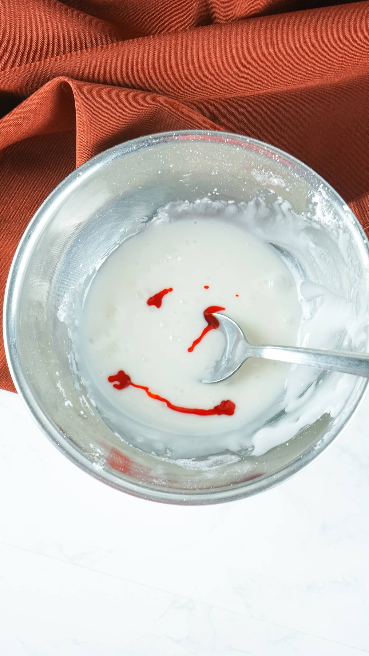 A metal bowl of white icing with red food coloring forming a smiley face, and a spoon inside—perfect for your next canned cinnamon roll hack or for adding flair to homemade pumpkin treats.