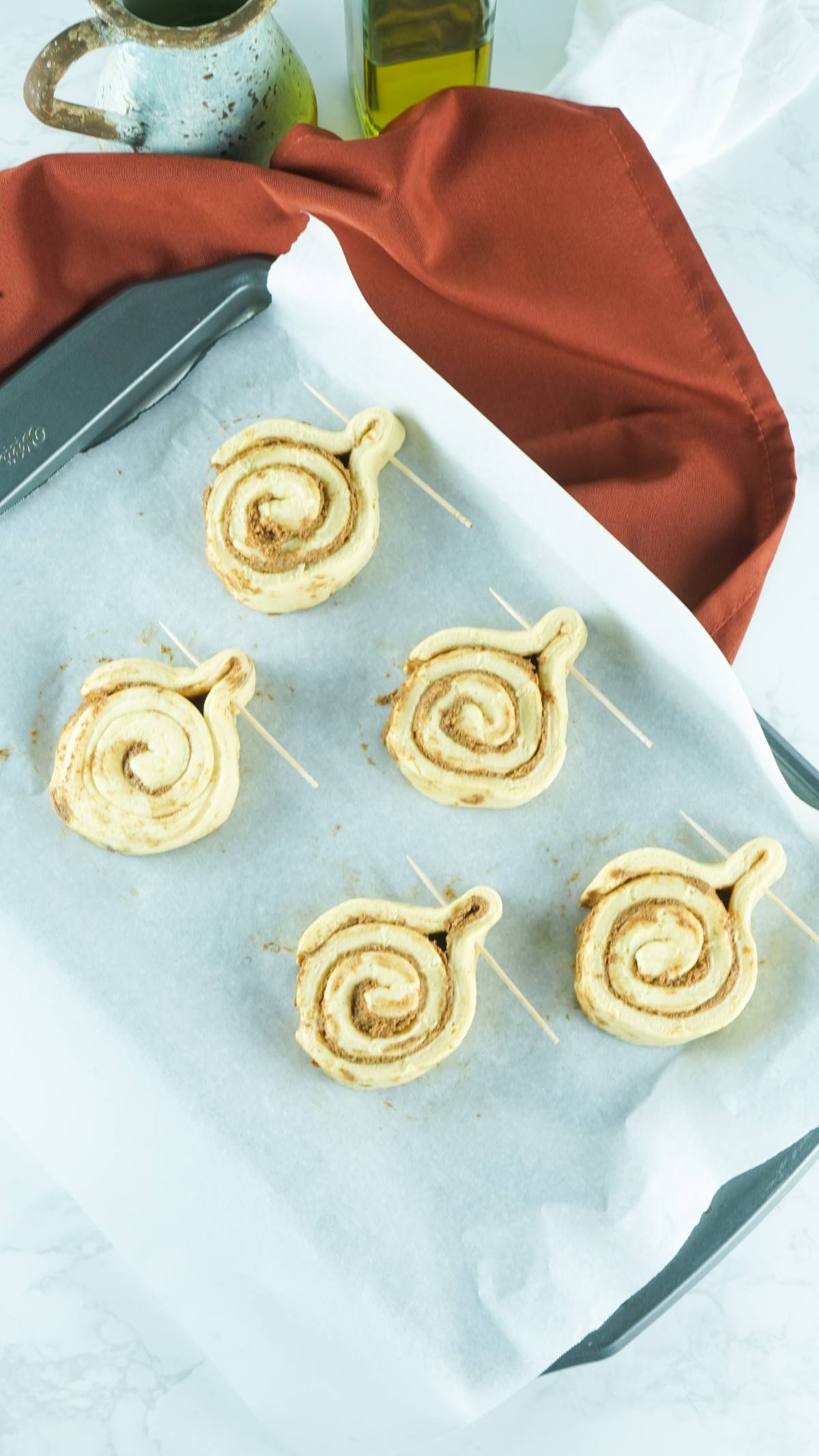 A baking sheet with five pumpkin shaped dessert ideas using canned cinnamon roll hack, arranged on sticks and ready to be baked on parchment paper.