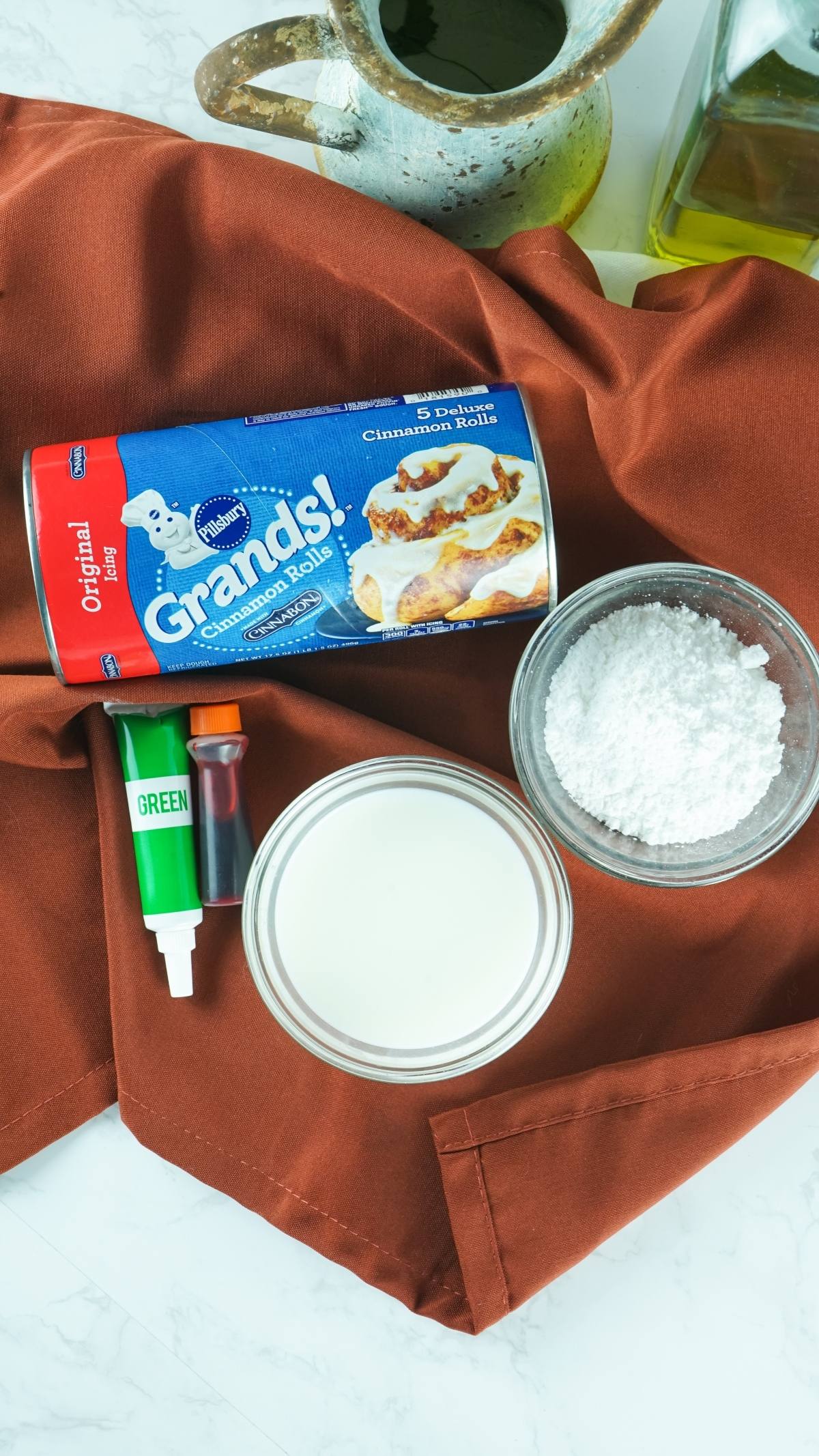 A can of cinnamon rolls, milk, powdered sugar, food coloring, and oil pitcher on a brown cloth—perfect for a pumpkin shaped dessert idea or an easy canned cinnamon roll hack.