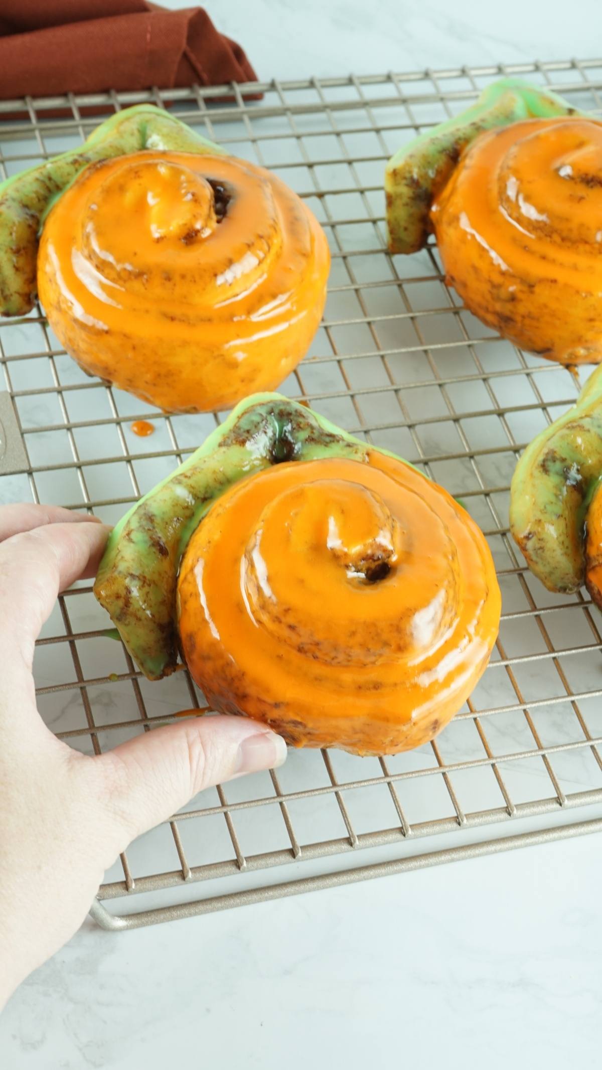 A hand reaches for a turtle-shaped pastry with orange icing—an adorable pumpkin shaped dessert idea inspired by Pumpkins made from Pillsbury Grand Cinnamon Rolls—resting on a cooling rack.