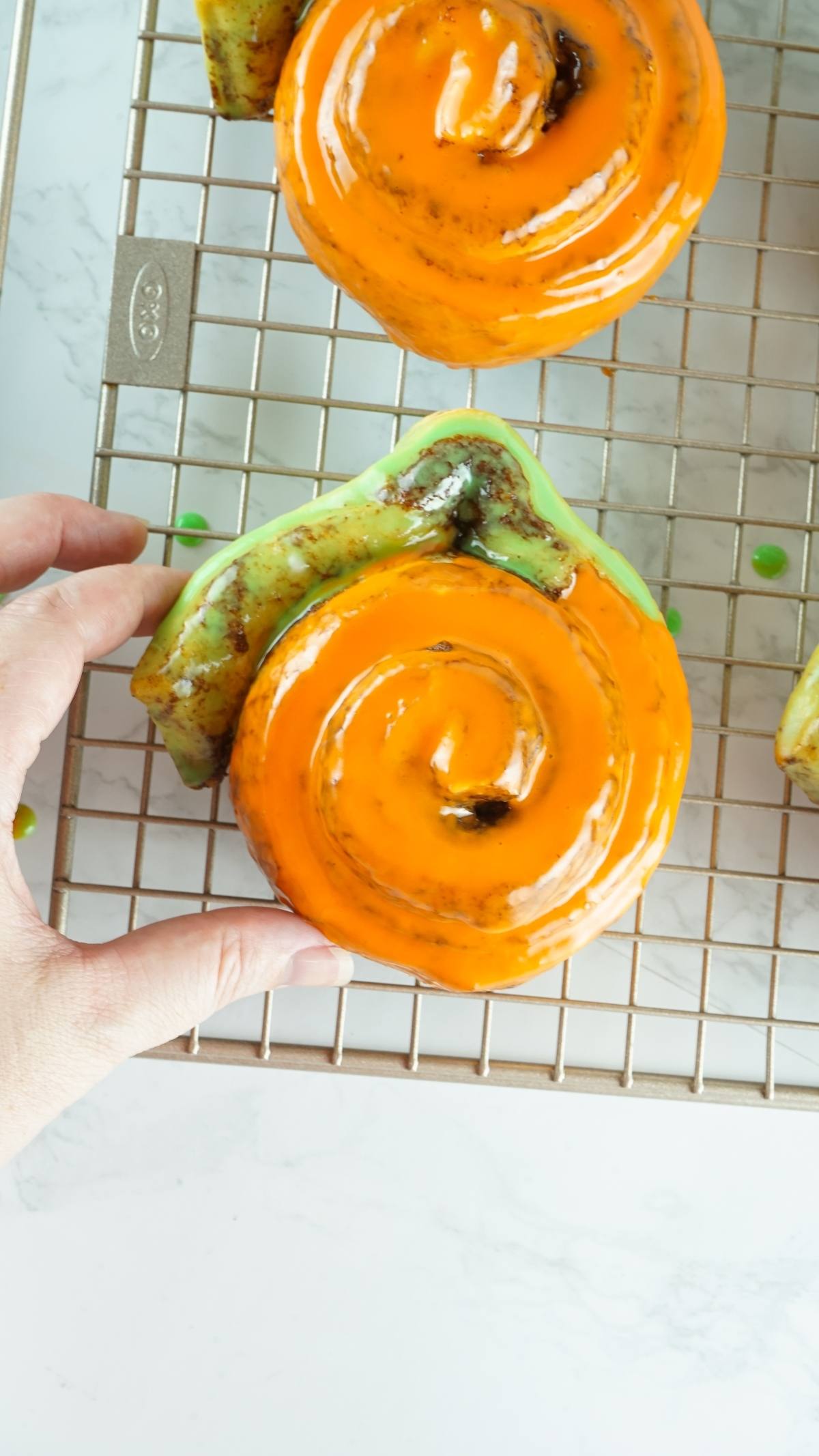 A hand holding an orange glazed cinnamon roll on a cooling rack, showcasing Pumpkin Shaped Cinnamon Rolls made from Pillsbury Grand Cinnamon Rolls—a delightful homemade pumpkin treat.