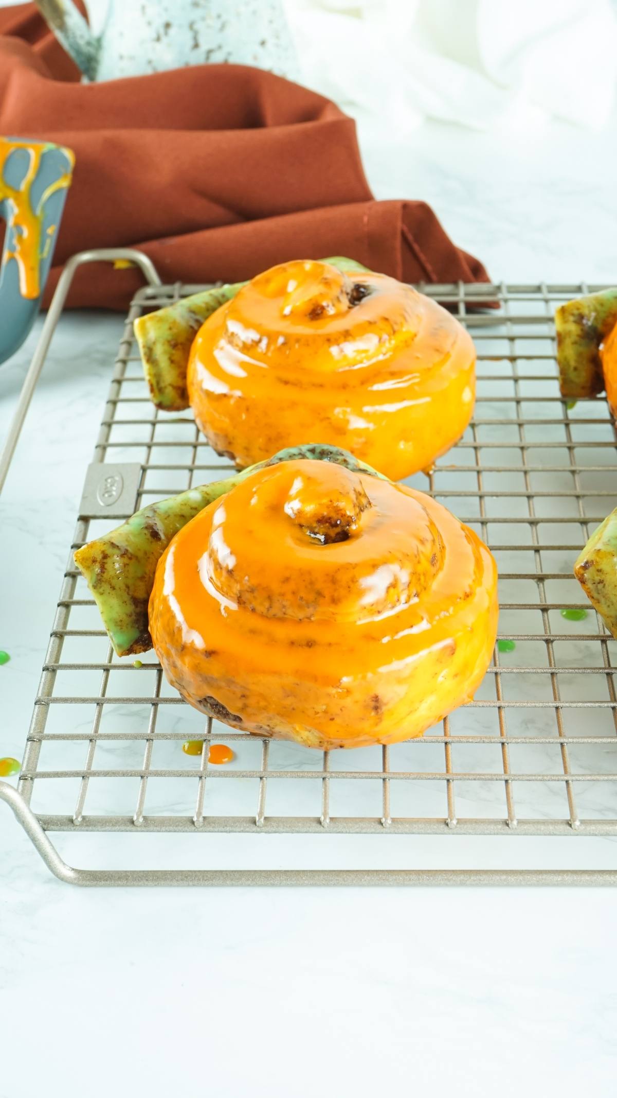 Three glazed Cinnamon rolls with bright orange icing, shaped like pumpkins, rest on a cooling rack—a perfect pumpkin shaped dessert idea for festive homemade pumpkin treats. A brown cloth provides the background.