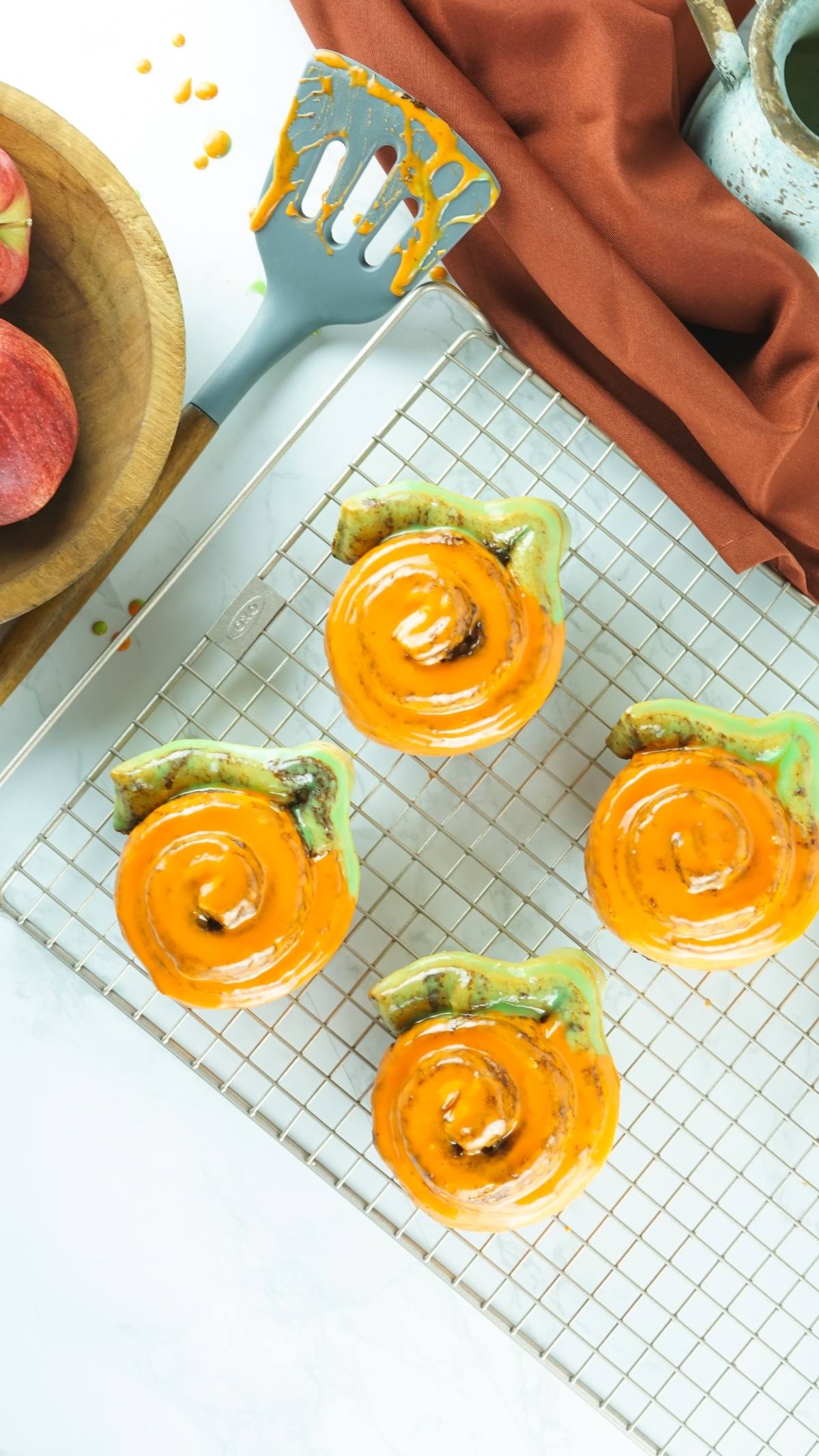 Four Pumpkin Shaped Cinnamon Rolls, glazed in orange and shaped like snails, cool on a rack with a spatula and apples nearby—delightful homemade pumpkin treats for autumn.