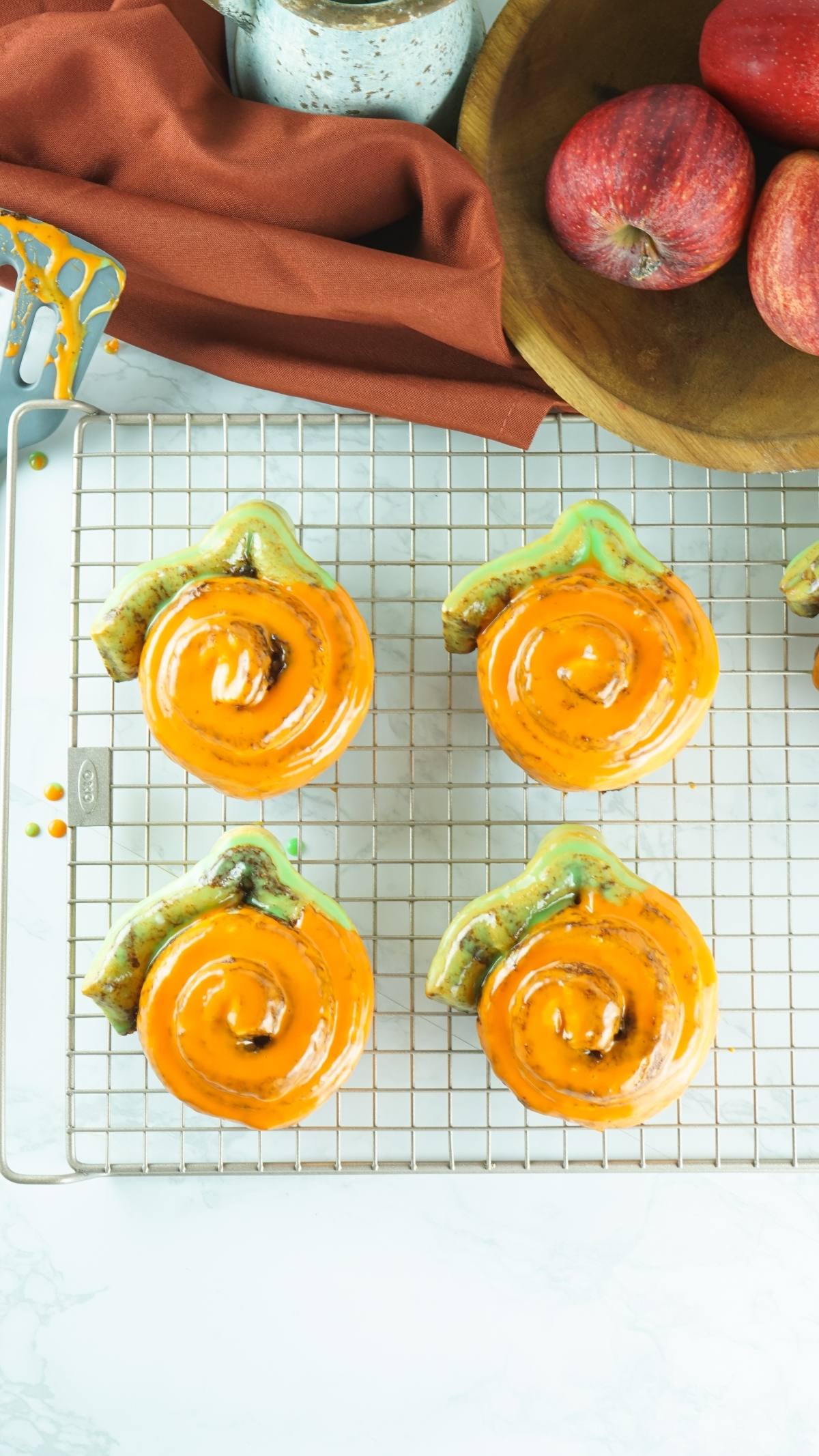 Four pumpkin-shaped cinnamon rolls with bright orange icing cool on a rack, next to red apples in a wooden bowl—a festive and easy pumpkin shaped dessert idea using Pillsbury Grand Cinnamon Rolls.