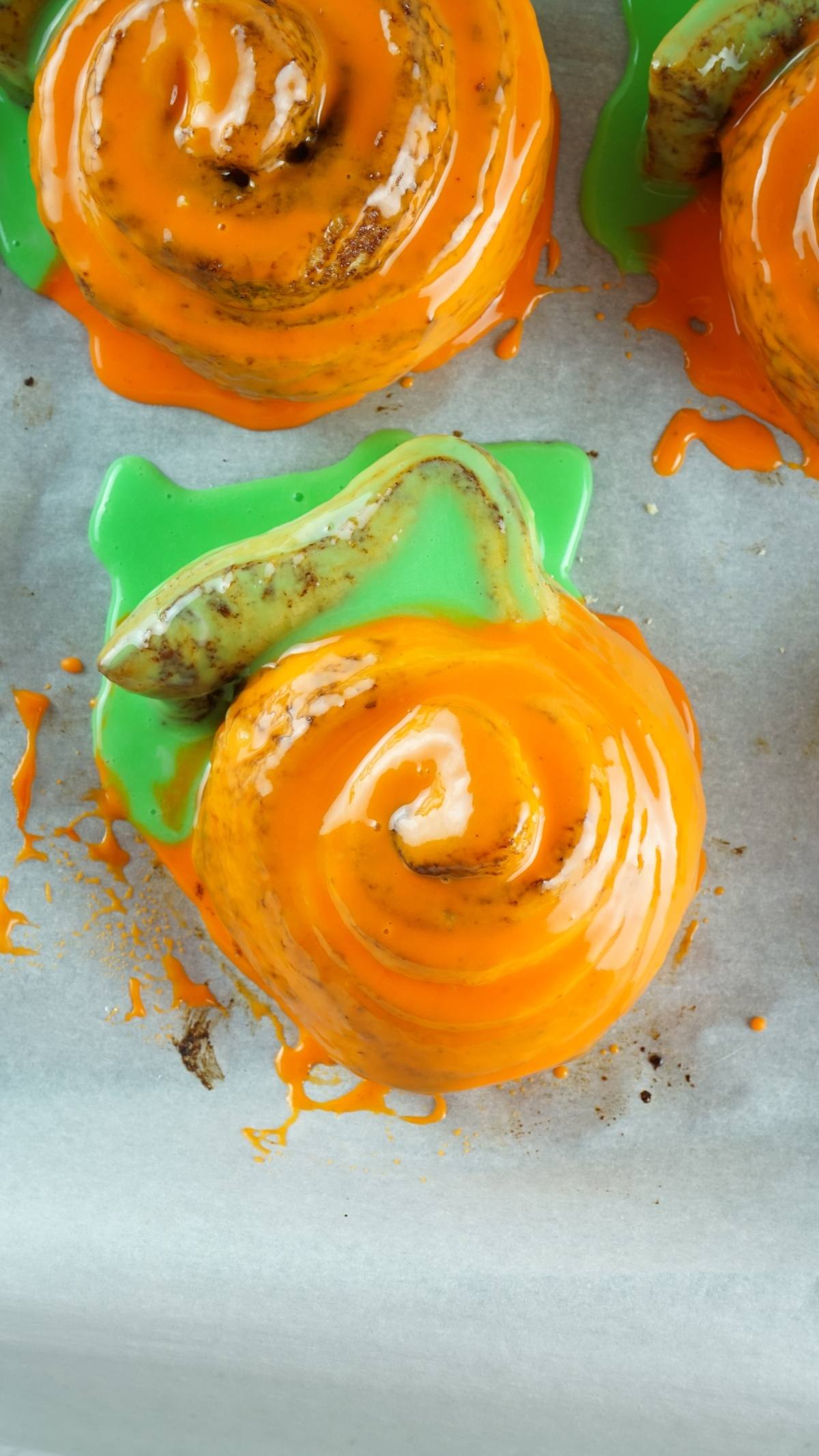 Pumpkin Shaped Cinnamon Rolls made from Pillsbury Grand Cinnamon Rolls, topped with orange and green icing to resemble pumpkins, are displayed on parchment paper.