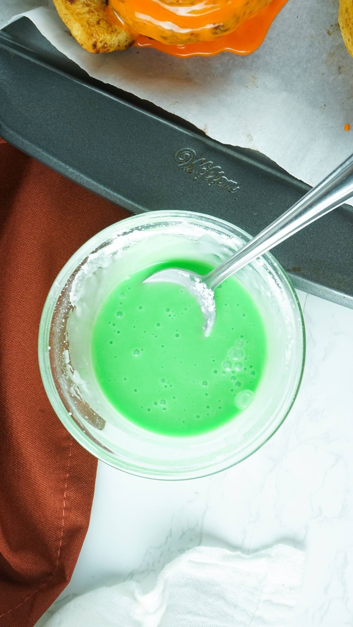 A glass bowl of green icing with a spoon sits next to a baking tray and brown cloth, ready for decorating Pumpkin Shaped Cinnamon Rolls.