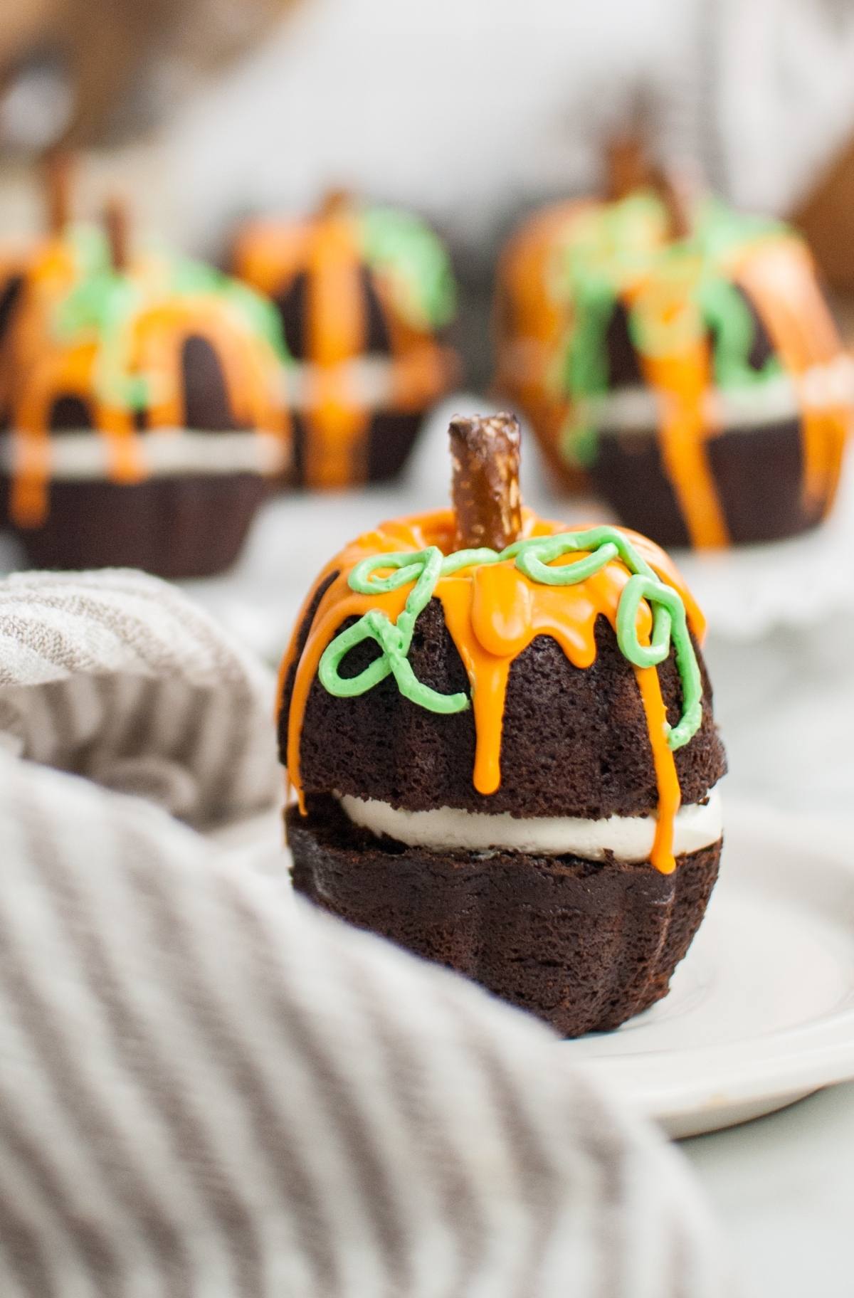 Chocolate cake shaped like a pumpkin, with orange and green icing and a pretzel stem, makes a festive Halloween pumpkin cake that’s perfect as a Thanksgiving or Halloween dessert. Served on a white plate.
