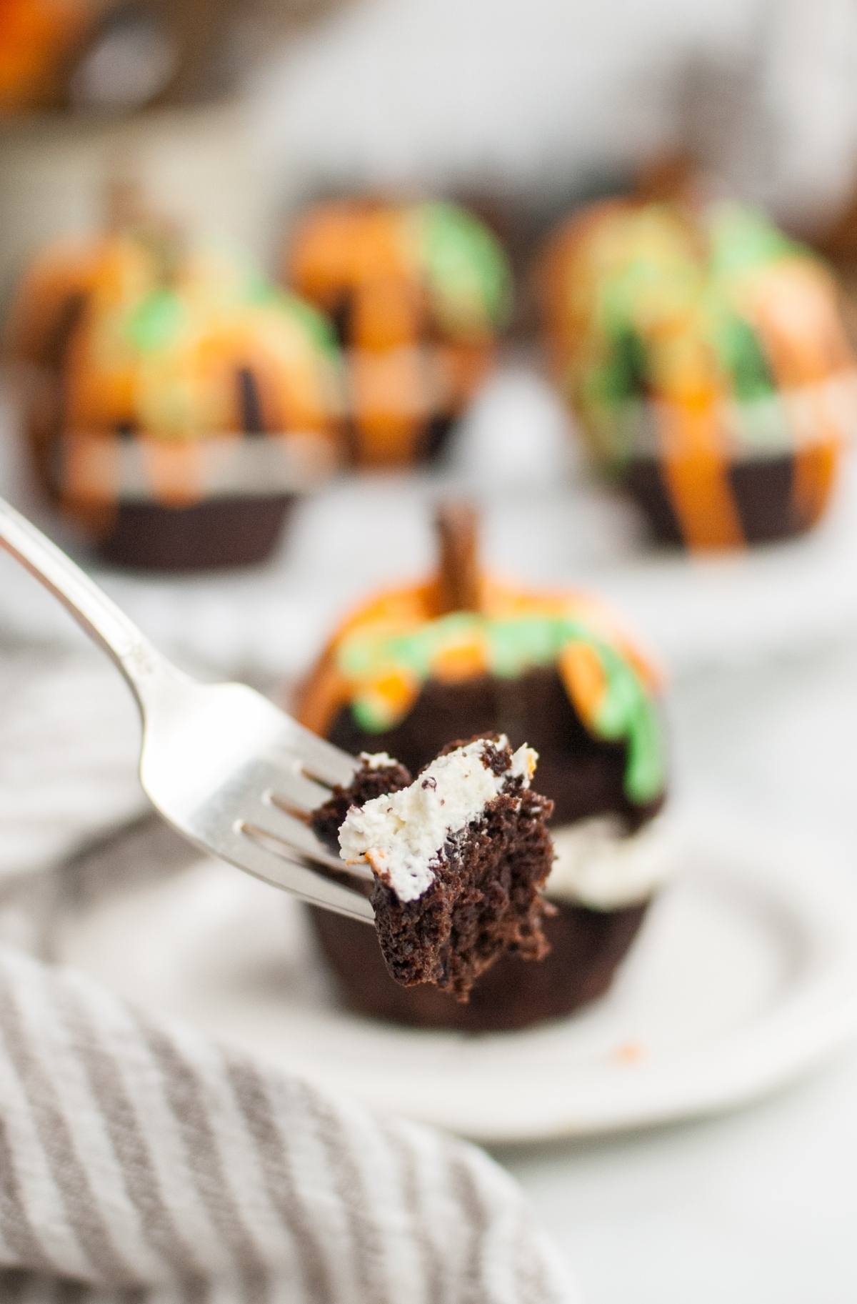 A fork holds a bite of chocolate cupcake with cream filling; in the background, decorated cupcakes and a pumpkin bundt cake set the scene.