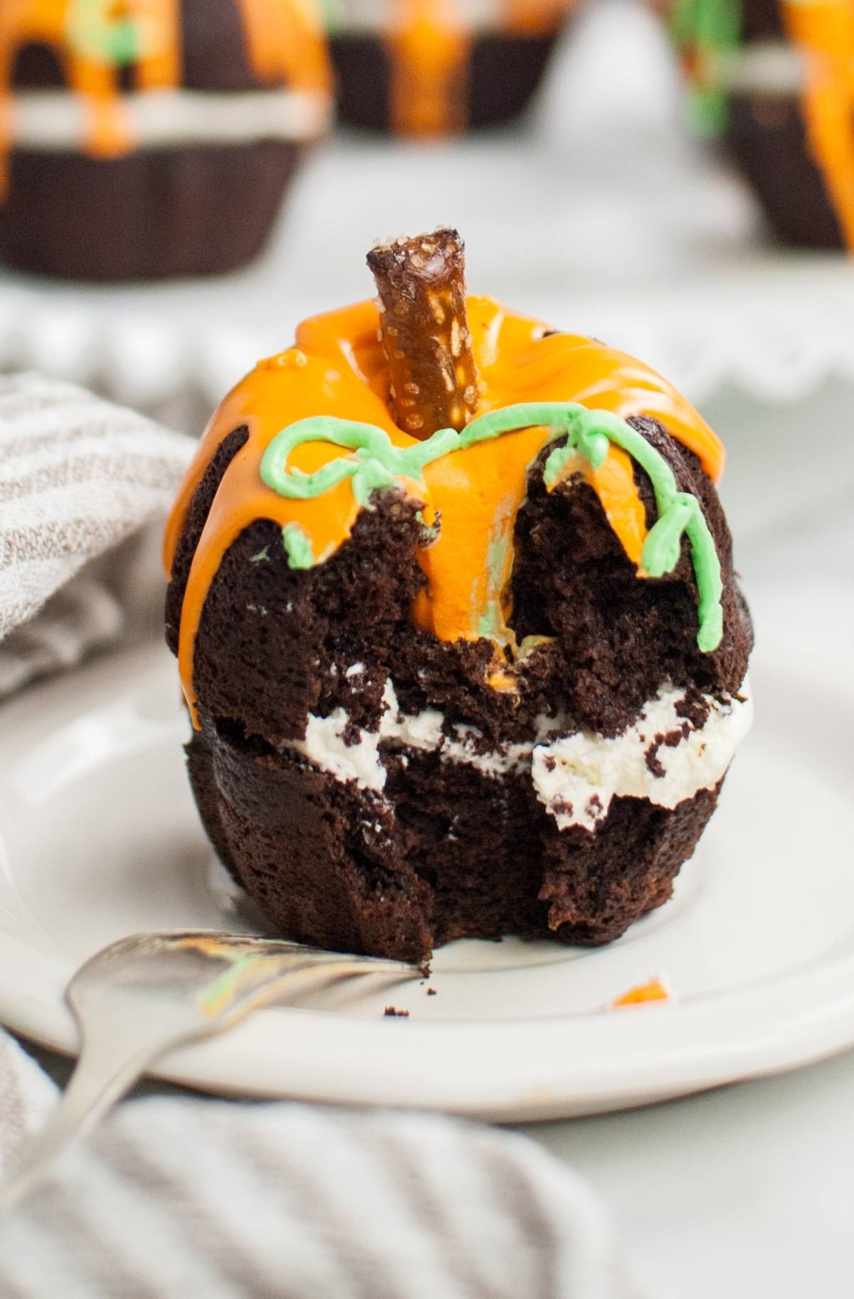 Chocolate cupcake shaped like a pumpkin with orange icing, creamy filling, and a pretzel stem, partially eaten—a playful twist on Halloween pumpkin cakes, perfect as a Thanksgiving or Halloween dessert.