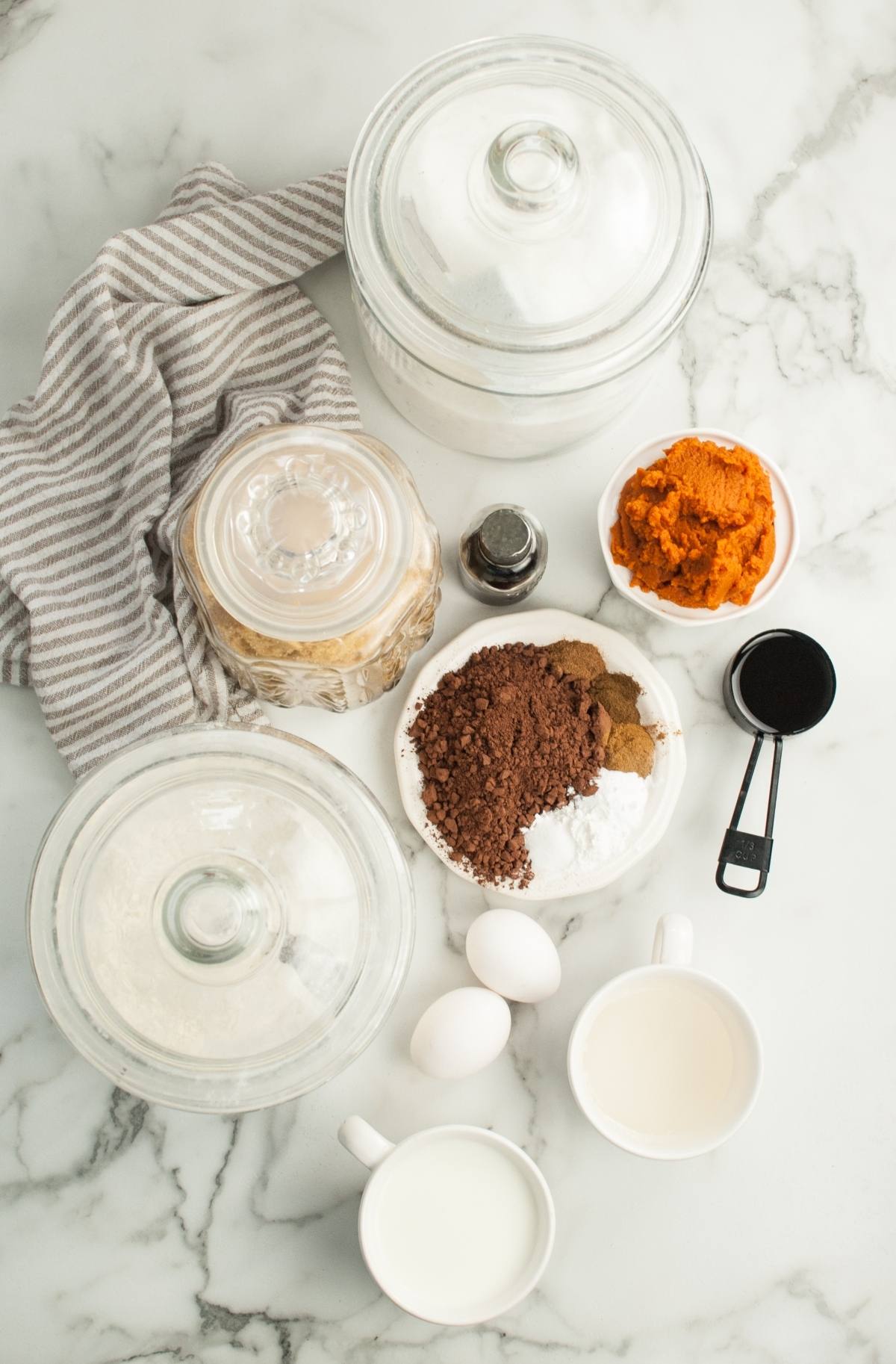 Baking ingredients on marble for Pumpkin Shaped Mini Bundt Cakes: eggs, cocoa, pumpkin puree, sugar, flour jars, vanilla, milk, and measuring cups—perfect for a festive Thanksgiving or Halloween dessert.