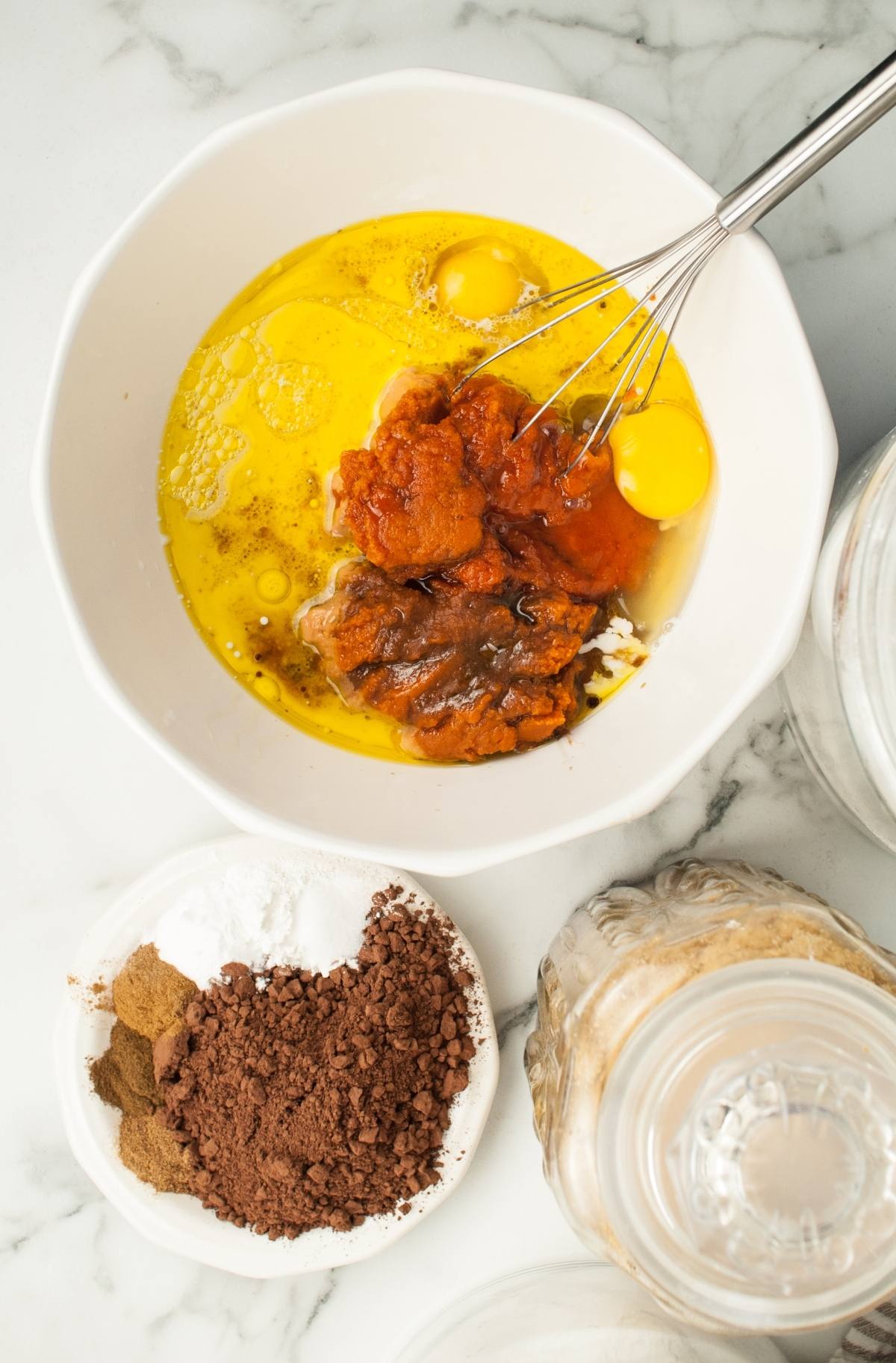 A bowl with eggs, pumpkin puree, and other ingredients being whisked, next to dishes of cocoa and spices—perfect for making a festive pumpkin bundt cake or Halloween pumpkin cakes.