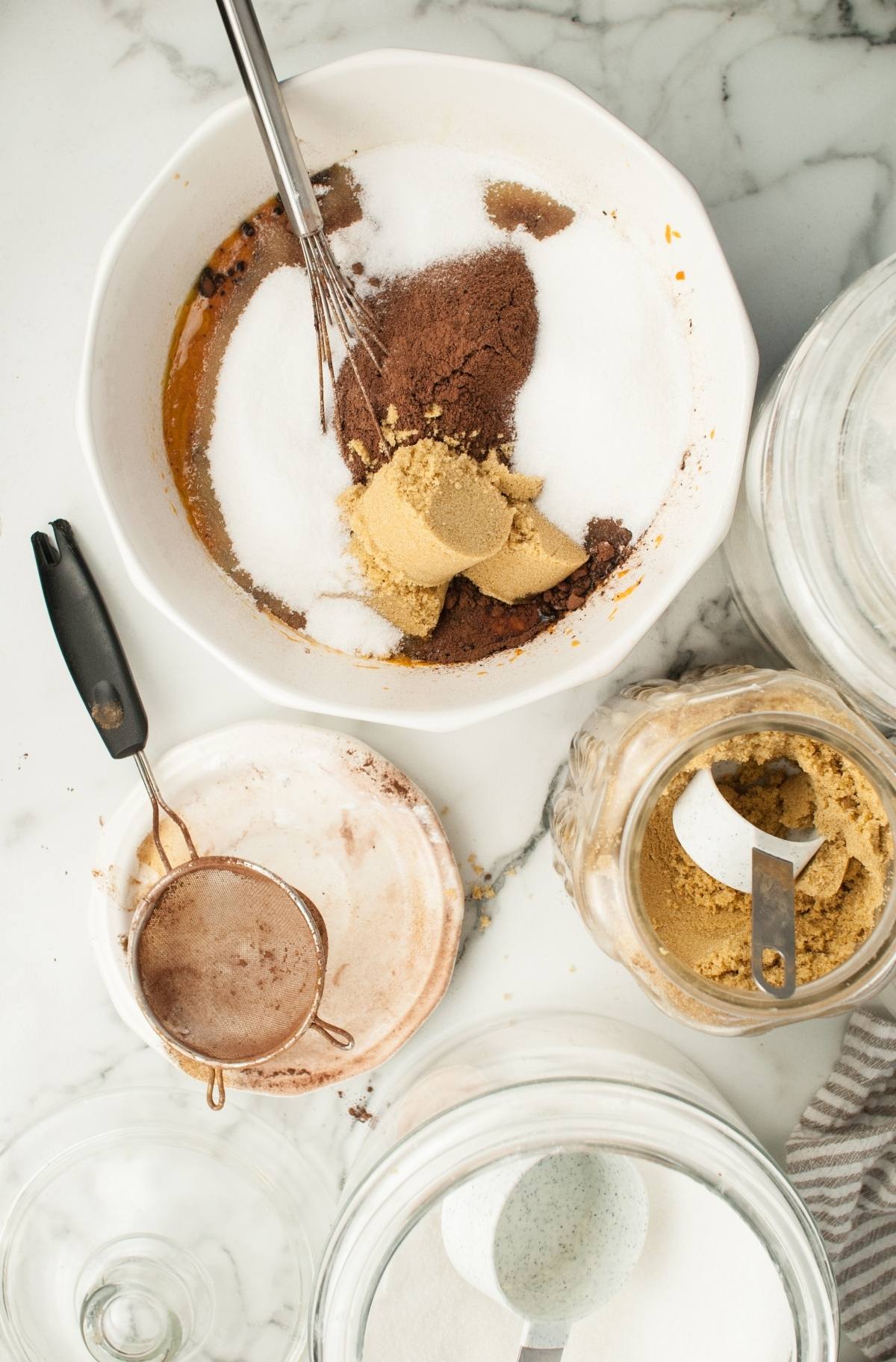 Baking ingredients in a bowl with a whisk; brown sugar, white sugar, and cocoa powder on a marble surface—perfect for making Pumpkin Shaped Mini Bundt Cakes as a festive Thanksgiving or Halloween dessert.