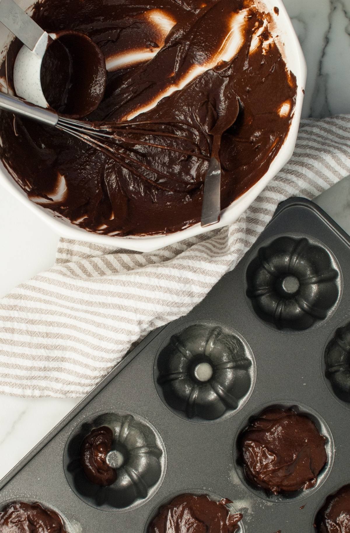 A bowl of chocolate batter with utensils sits beside a bundt cake pan and molds filled with batter, perfect for baking Pumpkin Shaped Mini Bundt Cakes as a festive Thanksgiving or Halloween dessert.
