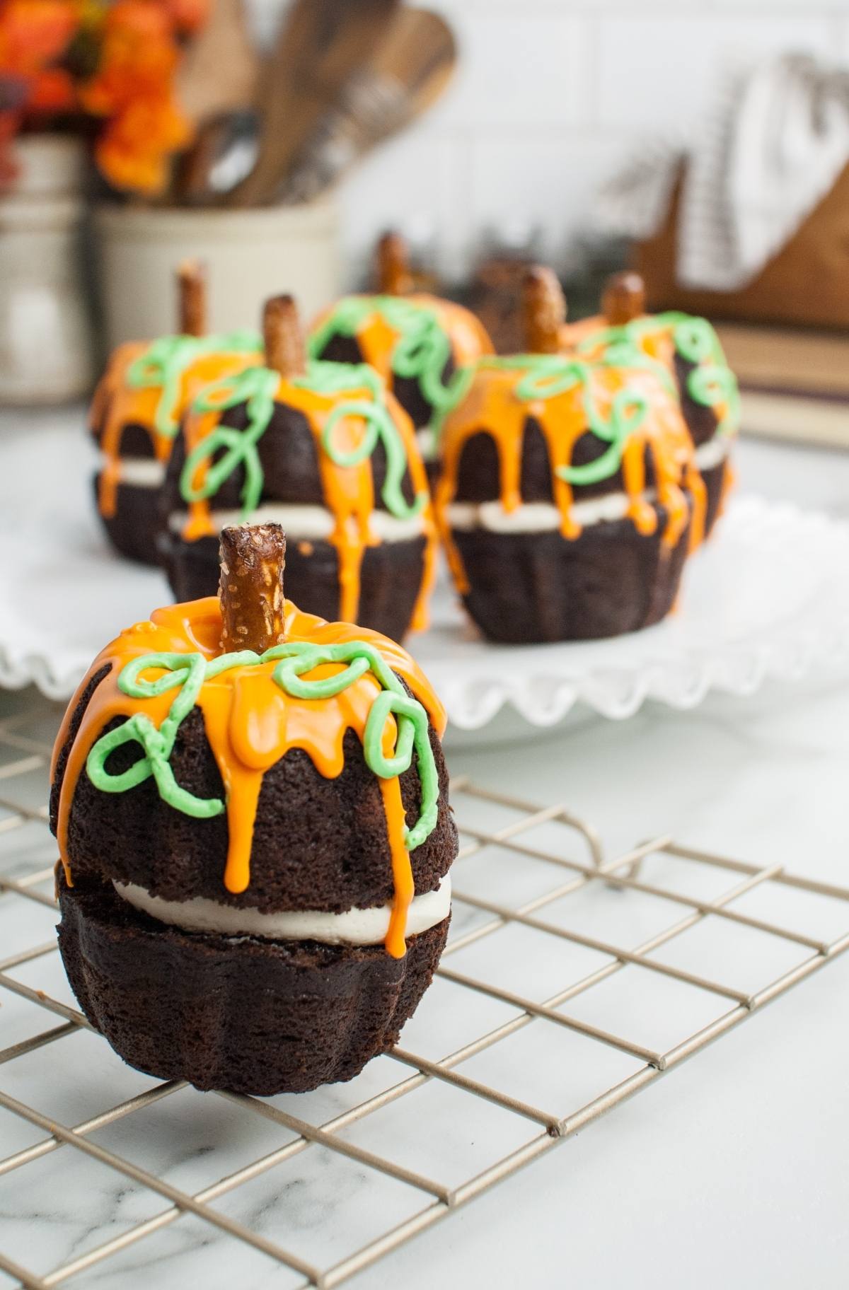 Pumpkin Shaped Mini Bundt Cakes with orange icing and pretzel stems rest on a cooling rack and serving plate—perfect as a festive Thanksgiving or Halloween dessert.