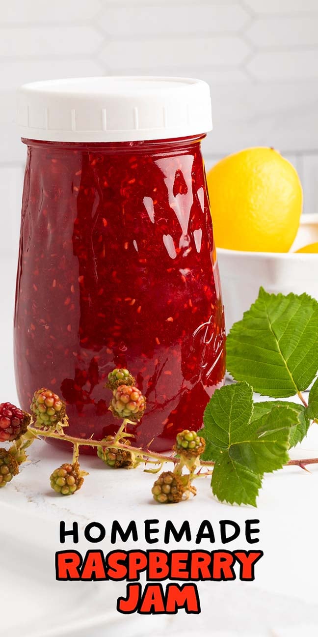A jar of homemade raspberry jam sits beside fresh raspberries, green leaves, and lemons on a white surface.