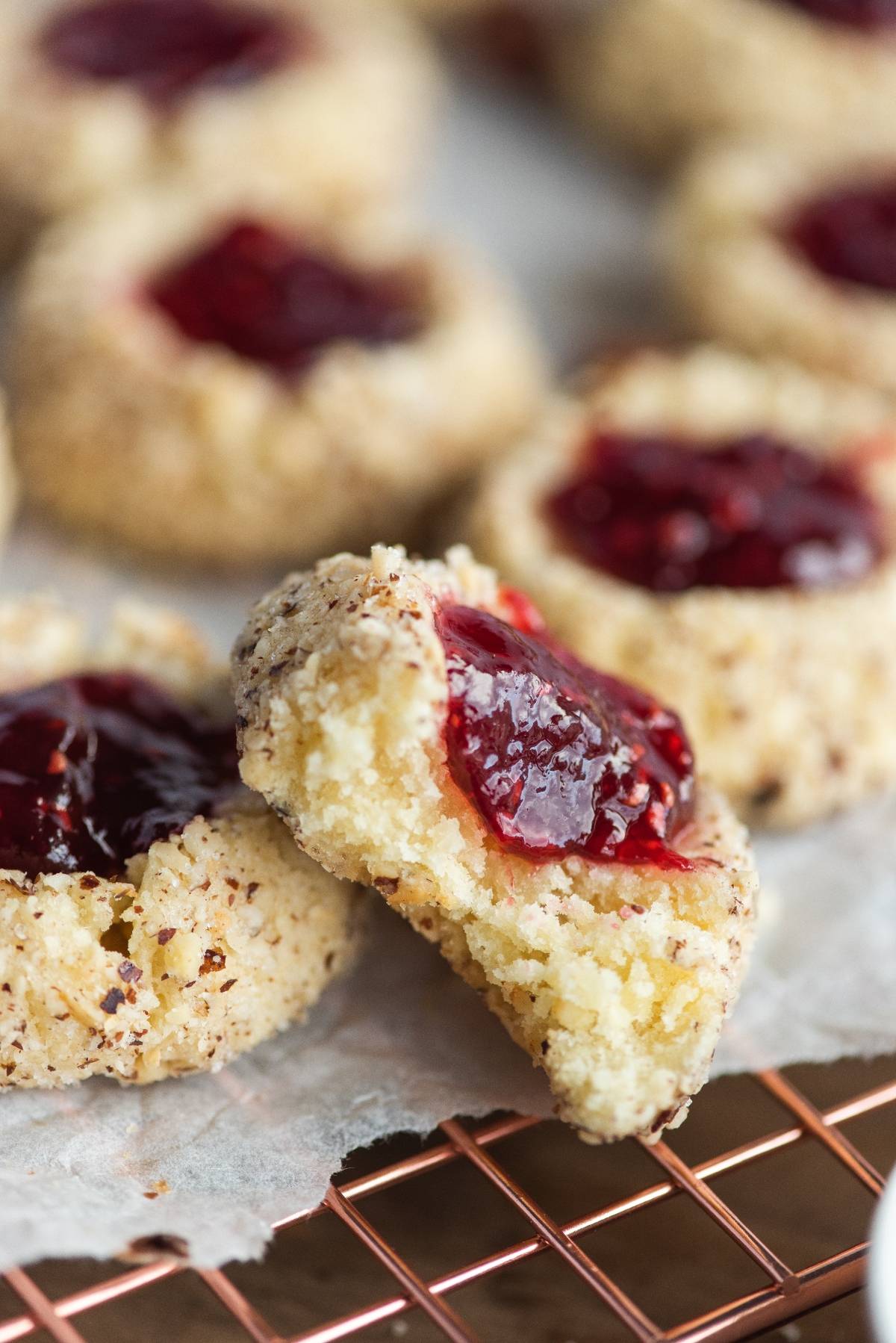 Close-up of raspberry hazelnut thumbprint cookies with vibrant red jam filling, one cookie broken in half to reveal the soft inside.