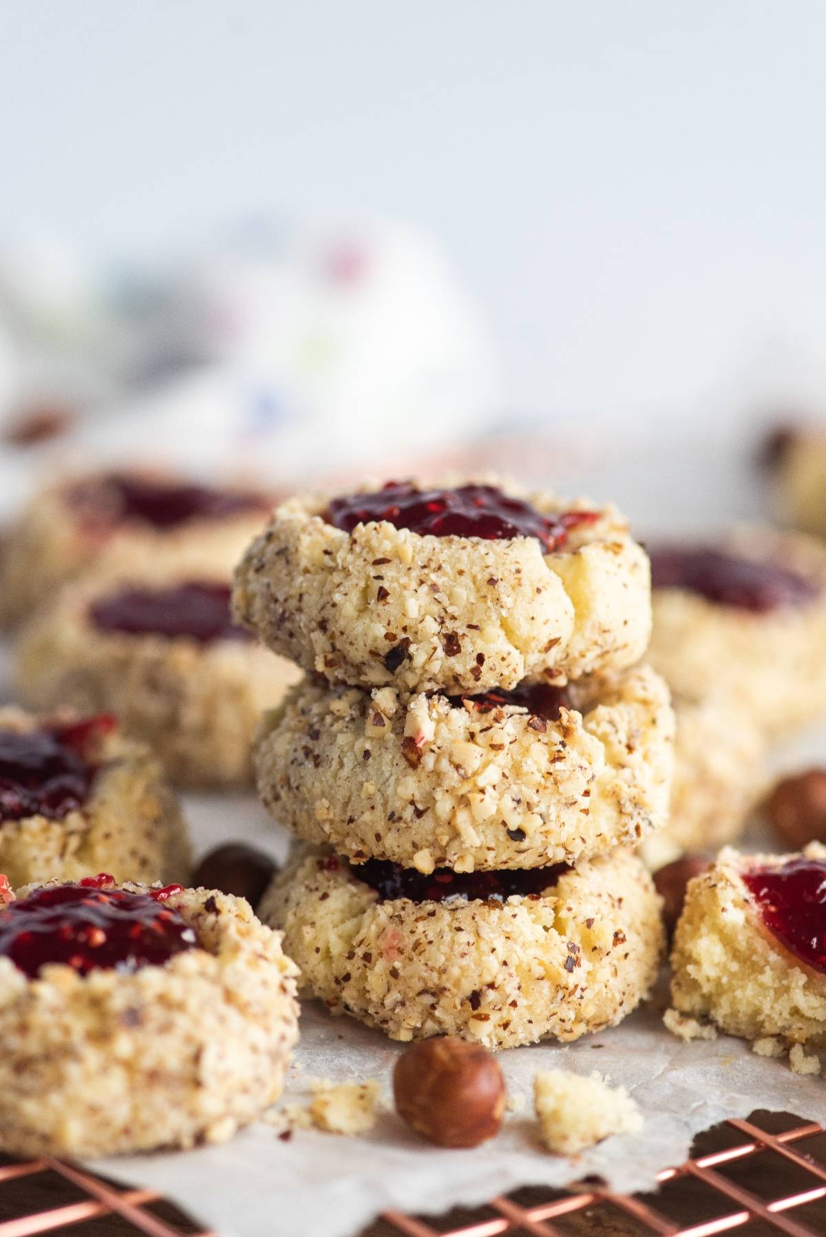 Three stacked classic thumbprint cookies with chopped nuts and red jam, surrounded by more hazelnut cookies and whole hazelnuts.